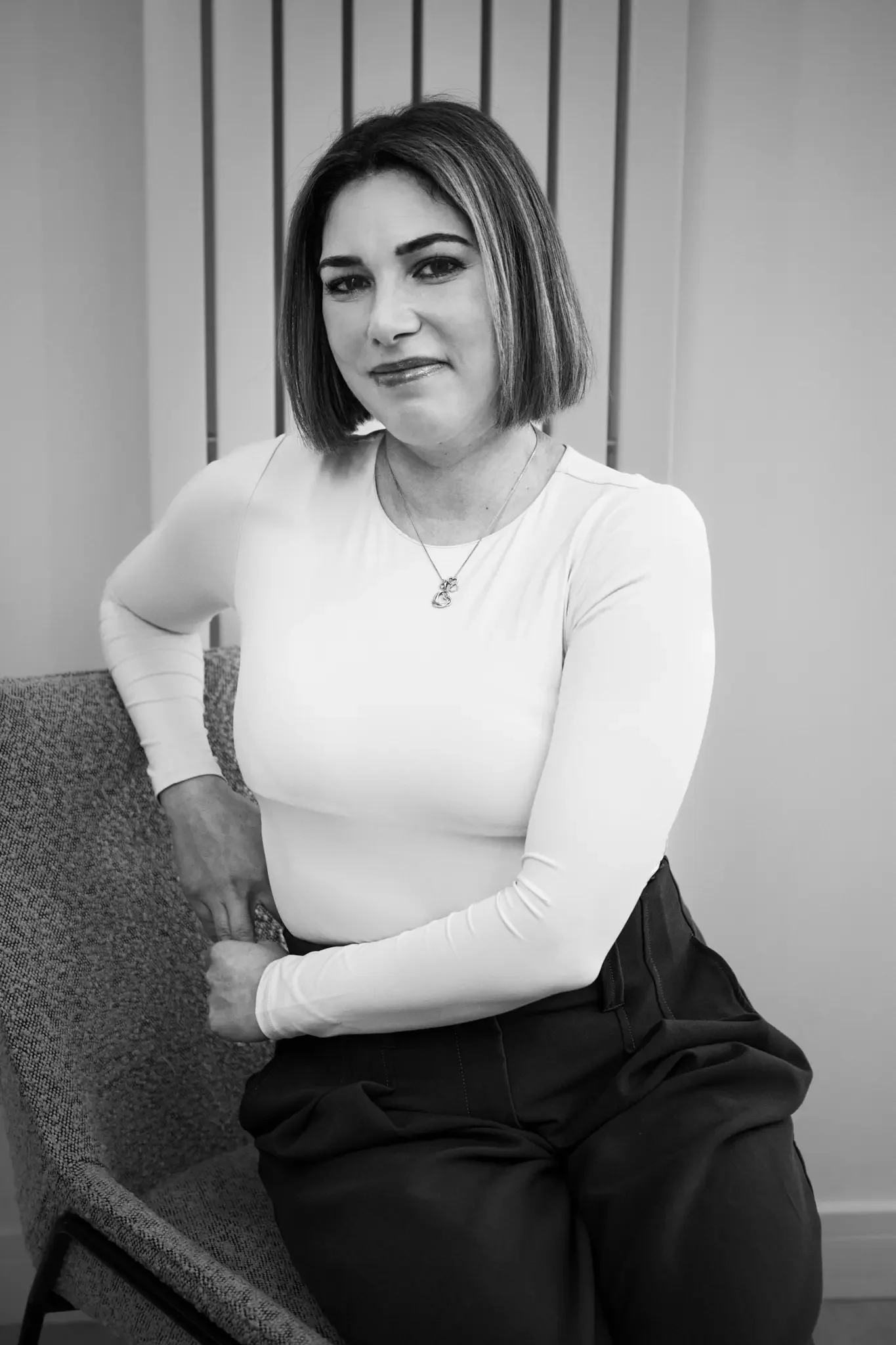 A woman with a shoulder-length bob hairstyle sitting on a chair with a high backrest, wearing a long-sleeved white top and dark pants, smiling slightly at the camera.
