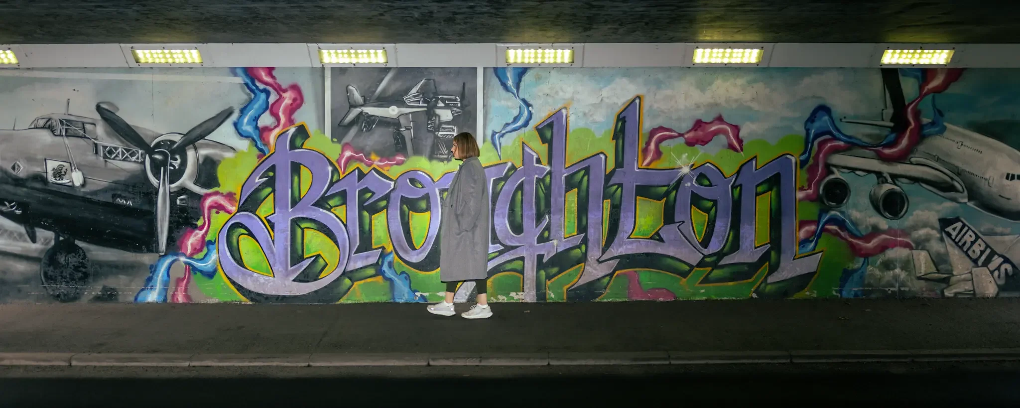 A person in a gray coat and white sneakers standing in front of colorful graffiti art of vintage airplanes and the words 'Berlin 11' on a wall.