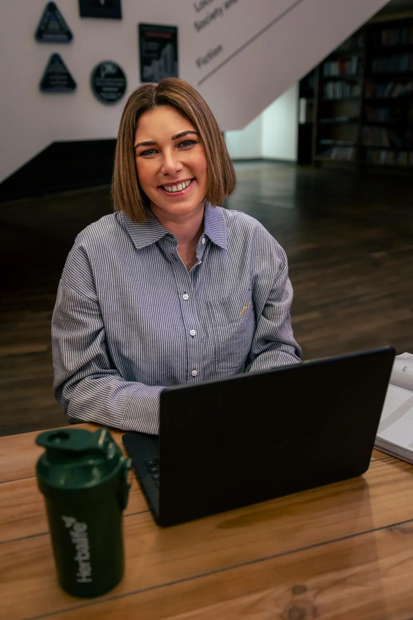 A woman with shoulder-length brown hair smiling while sitting at a wooden table with a black laptop and a green bottle in a modern, well-lit library or office space.