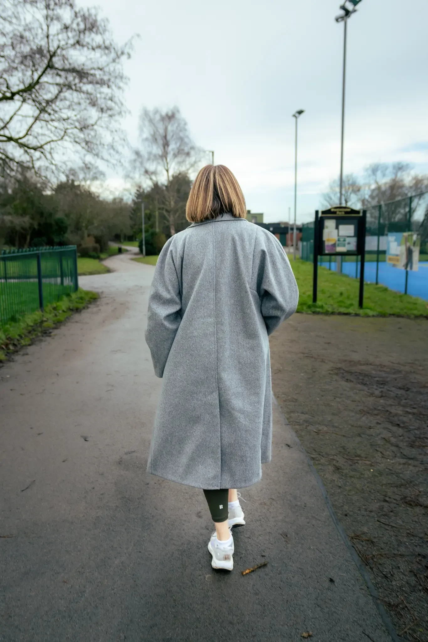 A woman in a gray coat and white sneakers walks along a park pathway with trees and a fenced sports area on either side, under a cloudy sky.