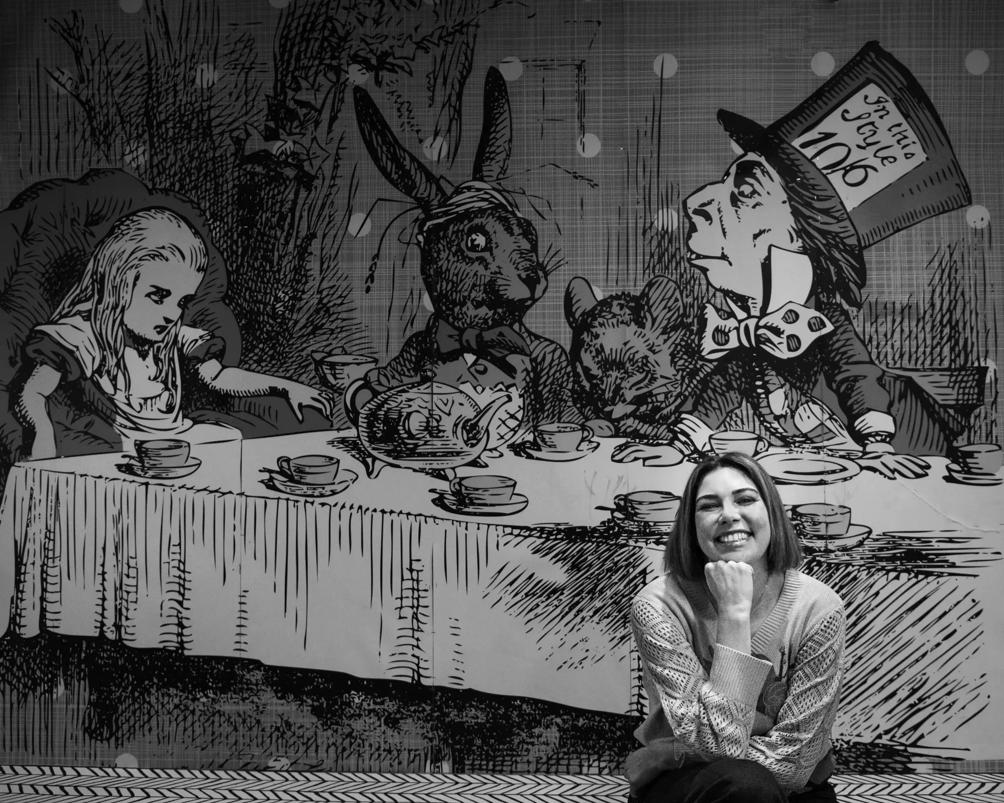 A woman smiling and sitting in front of a black and white illustration of a whimsical version of the last supper, featuring animals such as a rabbit, a cat, and a dog, with a large top hat on one of the characters that says "The Show 1906."