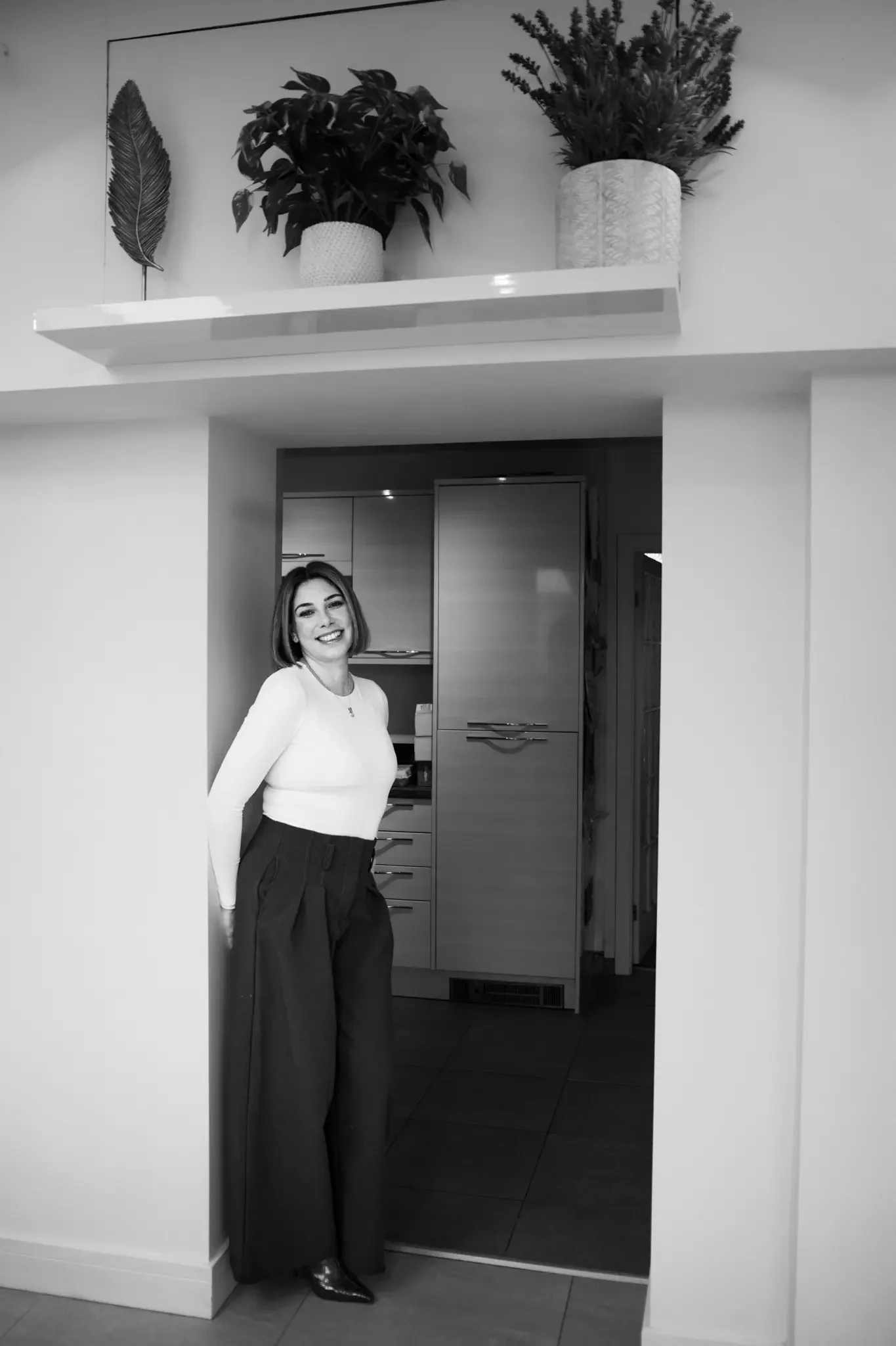 A smiling woman standing in a doorway in a home, with a kitchen visible behind her. The woman has short hair and is wearing a light-colored top and dark wide-leg pants. There is a decorative shelf above with potted plants and a feather ornament.