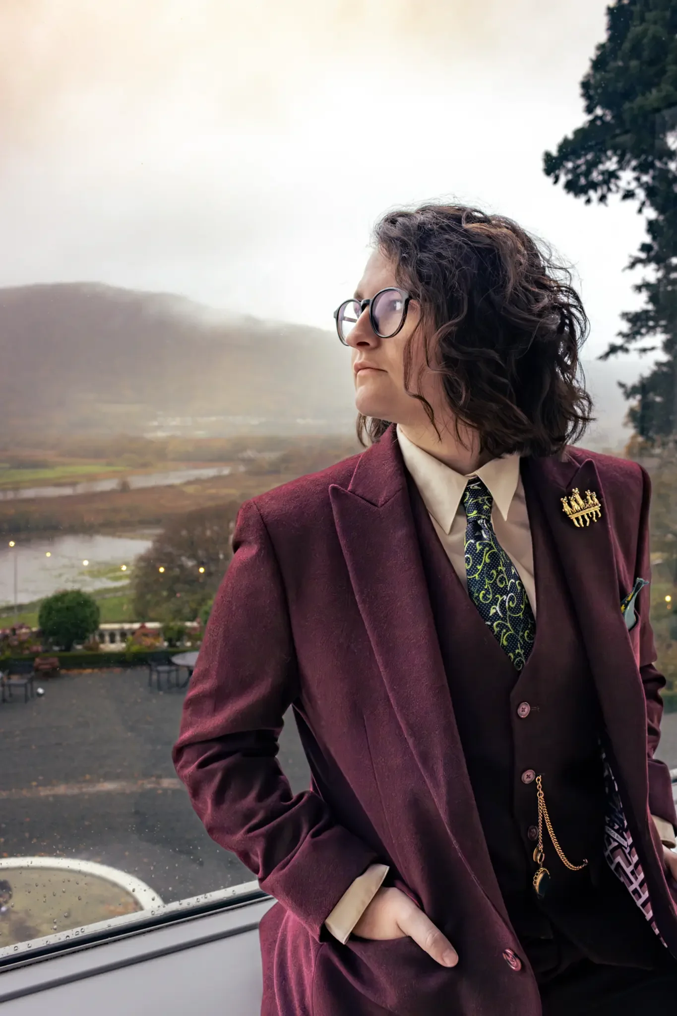 north wales wedding photographer- image of a bride in a burgundy tweed suit staring out the window on her wedding day at a north wales venue.webp