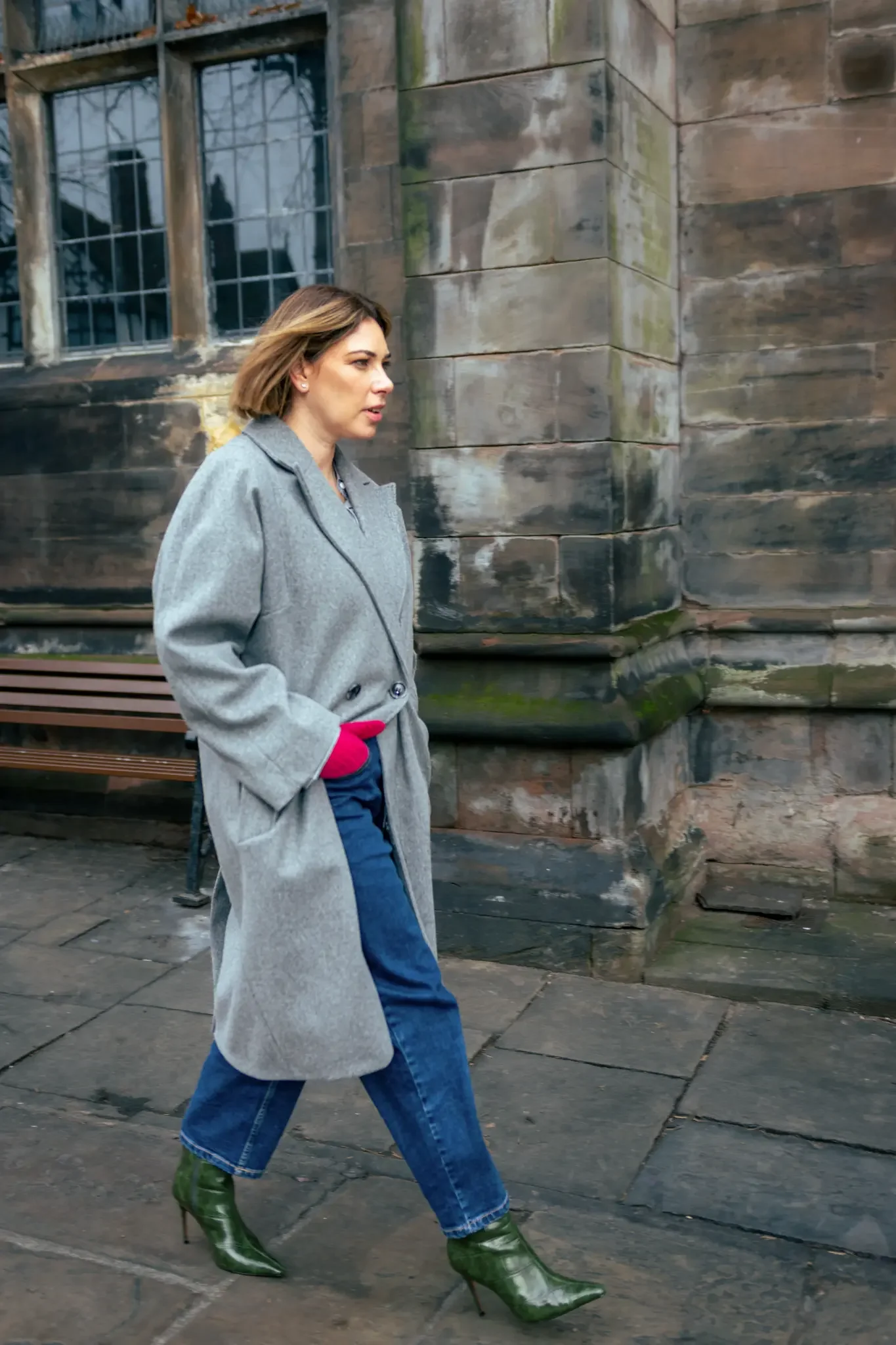 A woman walking on a stone sidewalk near a stone building, wearing a gray coat, blue jeans, green high-heeled boots, and red gloves.