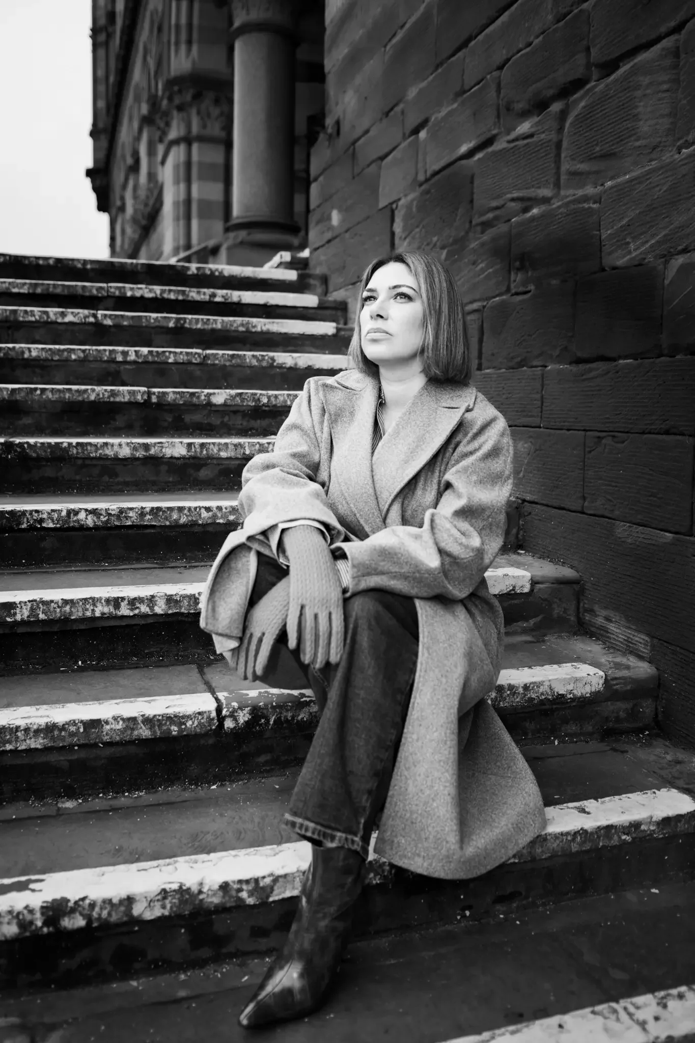 A woman sitting on outdoor stairs near a brick wall, looking thoughtfully into the distance, in black and white.