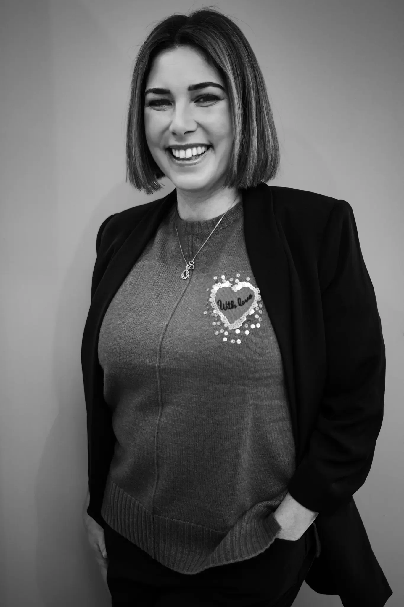 Smiling woman with shoulder-length hair wearing a dark blazer over a sweater with a heart-shaped decoration and the words "With Love".