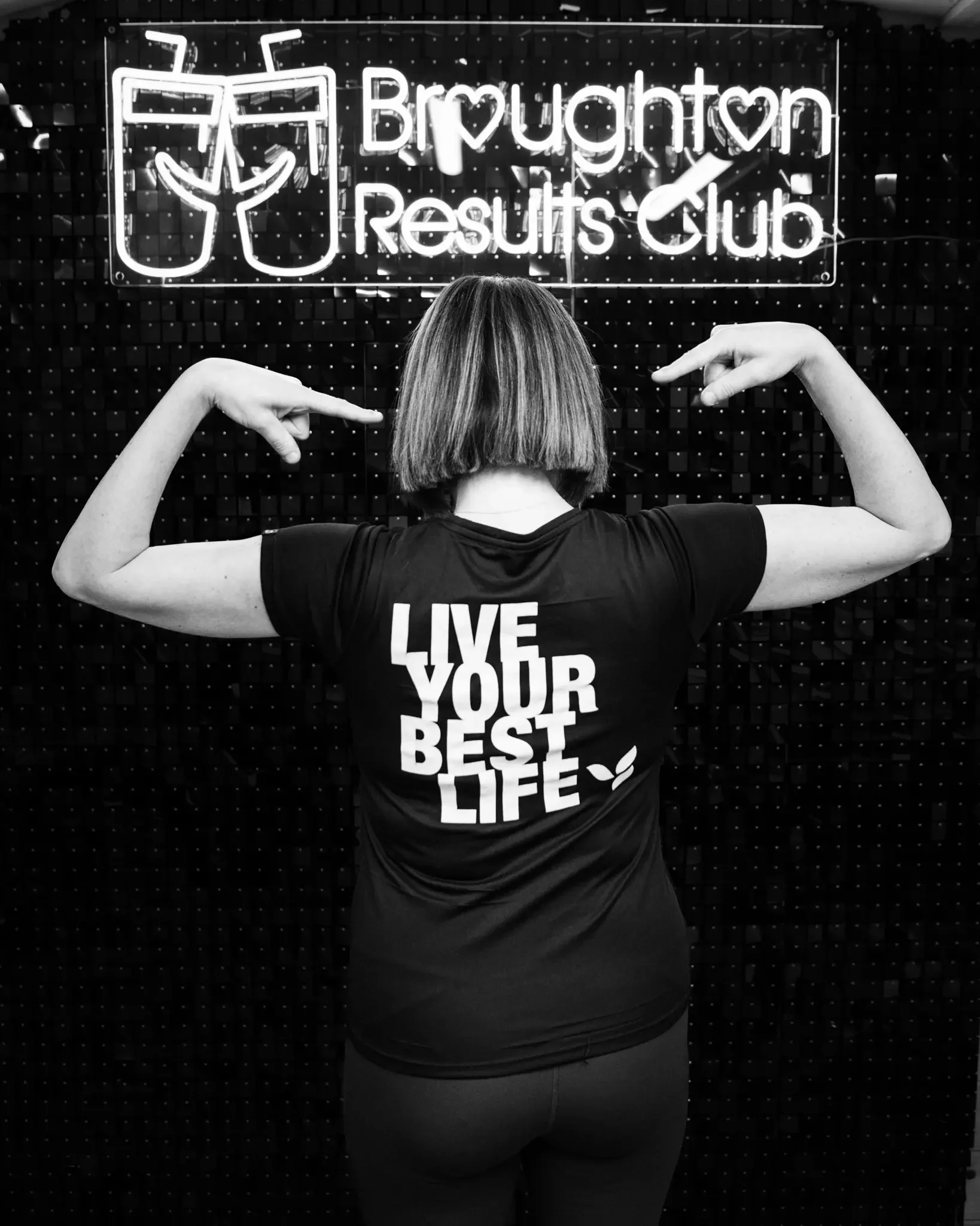 A person with short hair flexing their arm muscles in front of a sign that reads 'Brughton Results Club' with a cartoon of a trophy and a checkmark. The person is wearing a T-shirt that says 'LIVE YOUR BEST LIFE' with a small graphic of a leaf.