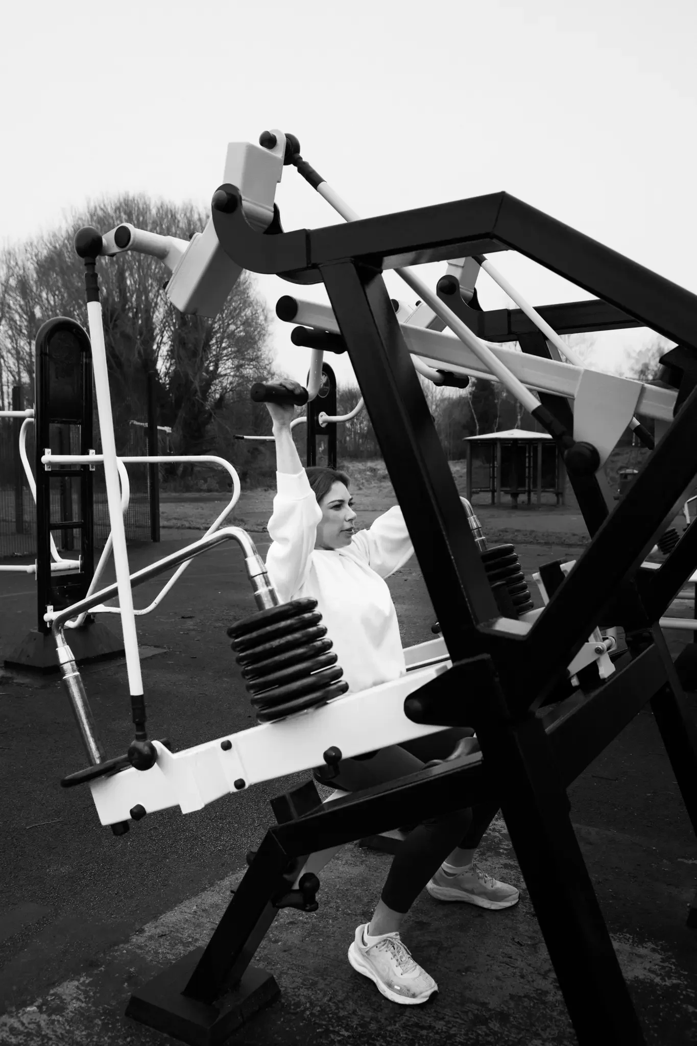A woman using outdoor gym equipment, lifting a bar overhead, in a park with trees in the background.