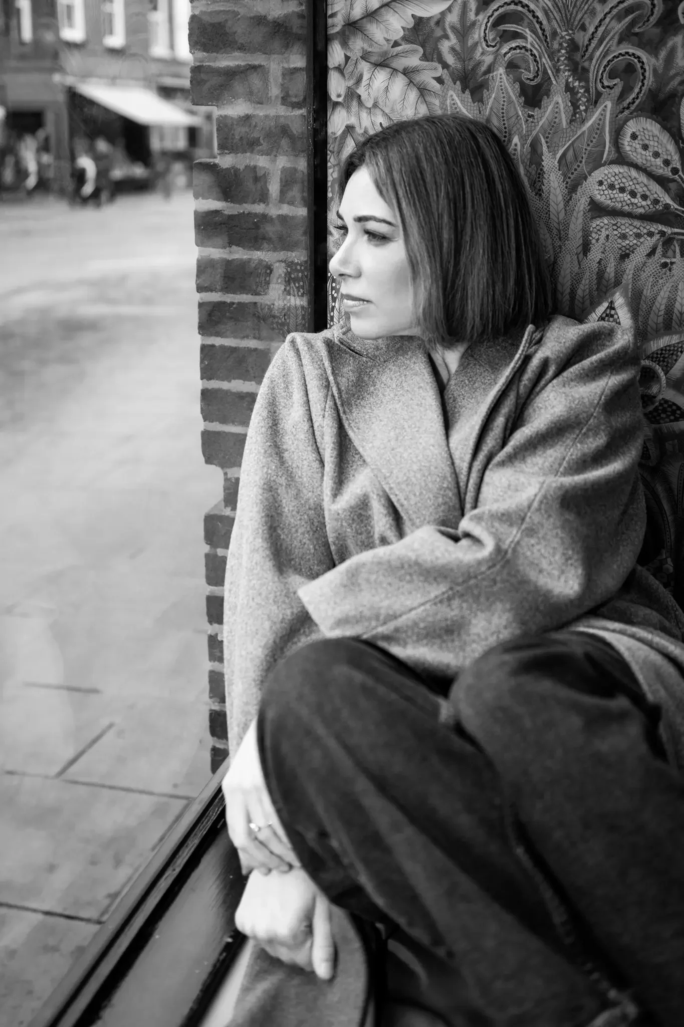 A woman with short hair sitting by a window, wearing a coat and looking outside with a contemplative expression.