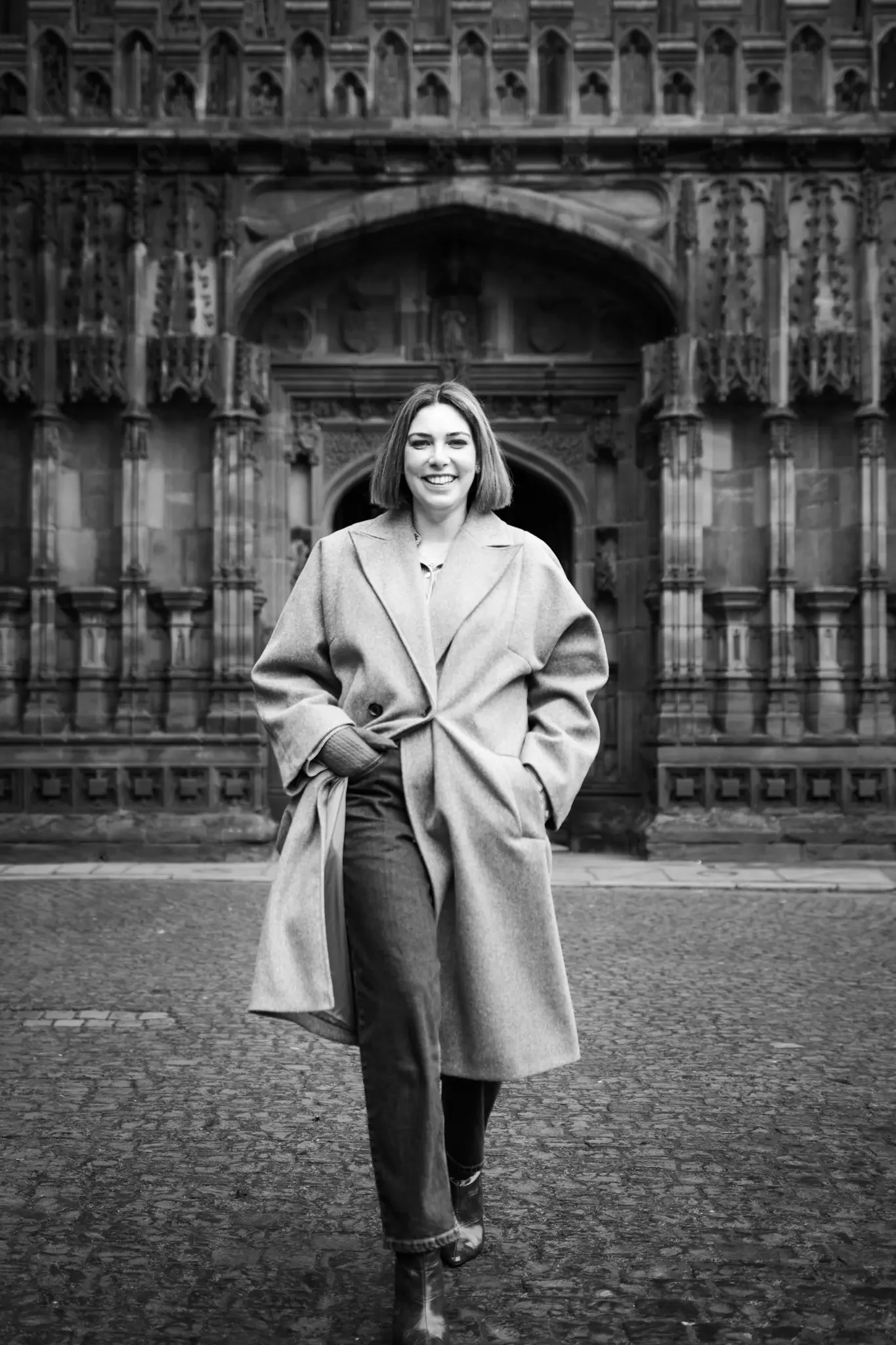 A woman with shoulder-length hair, smiling, standing in front of an ornate historic building, wearing a long coat, pants, and boots.