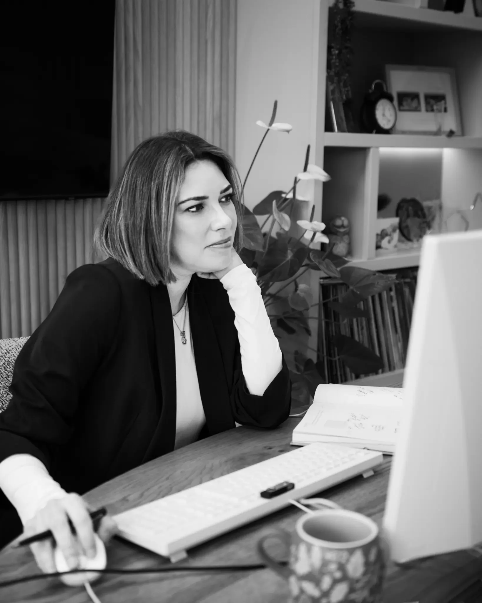 A woman in professional attire working on a computer at her desk, with an open notebook, a mug, and a computer mouse in front of her, in a home office setting.