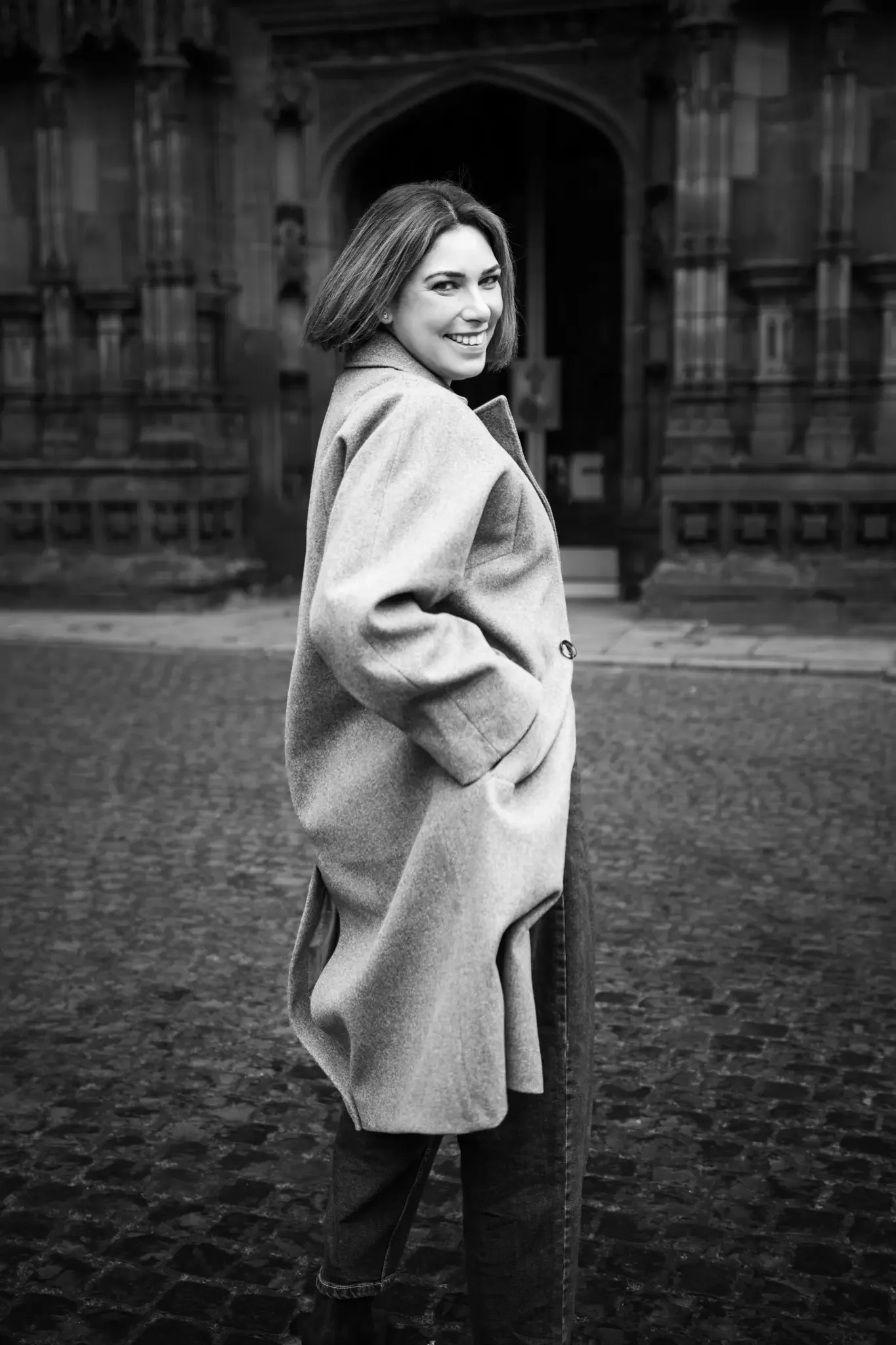 A woman smiling and walking on a cobblestone street in front of a historic building, dressed in a coat and jeans, in black and white photography.