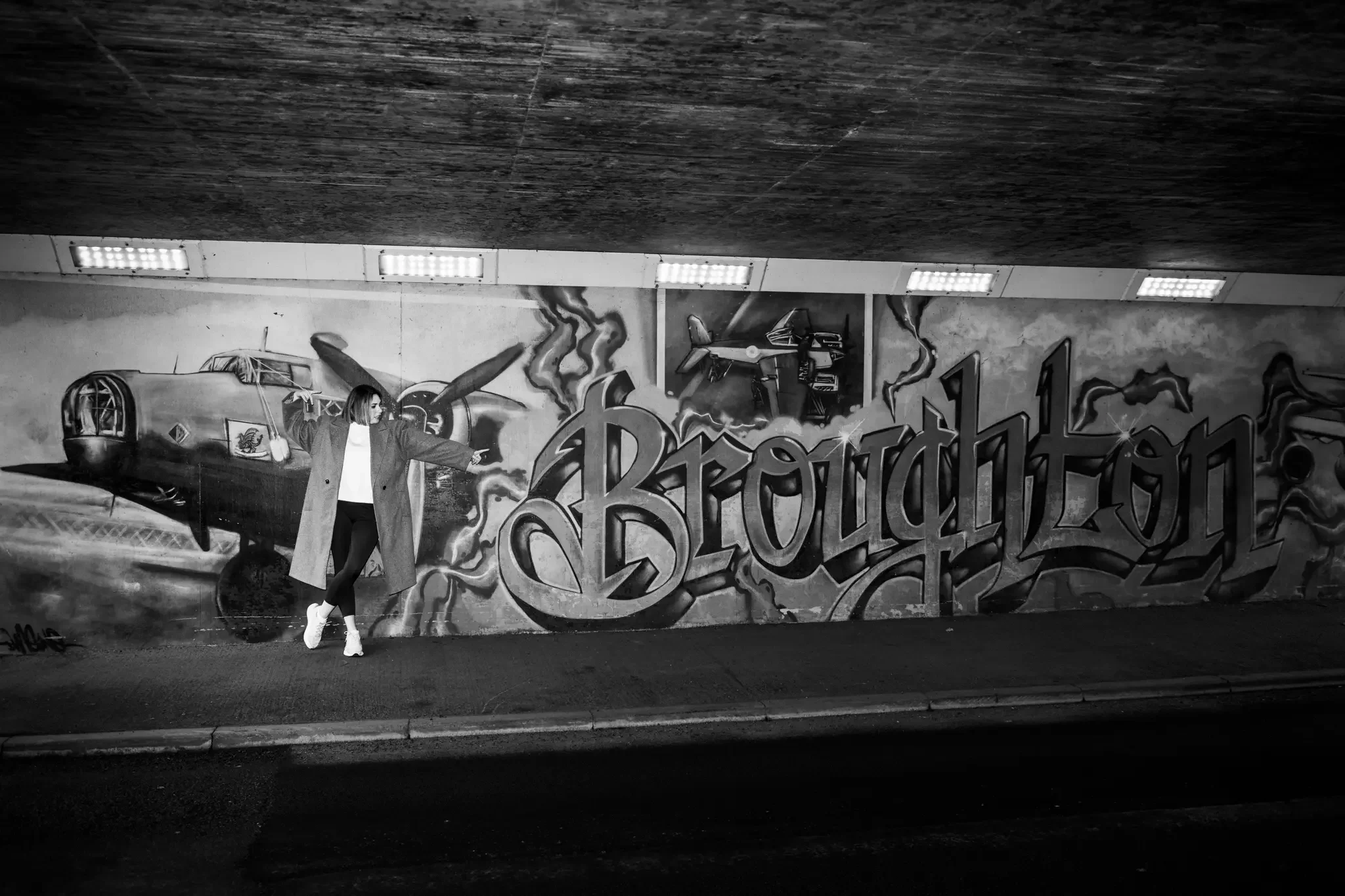 A person pointing towards a graffiti mural of vintage airplanes and the word 'Broughon' on a wall in an underpass. The mural is in black and white.