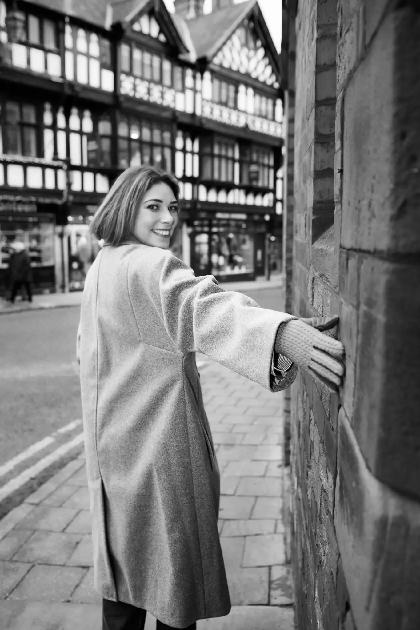 A woman smiling and looking back at the camera while leaning against a brick wall on a city sidewalk, wearing a coat and gloves, with modern buildings in the background.