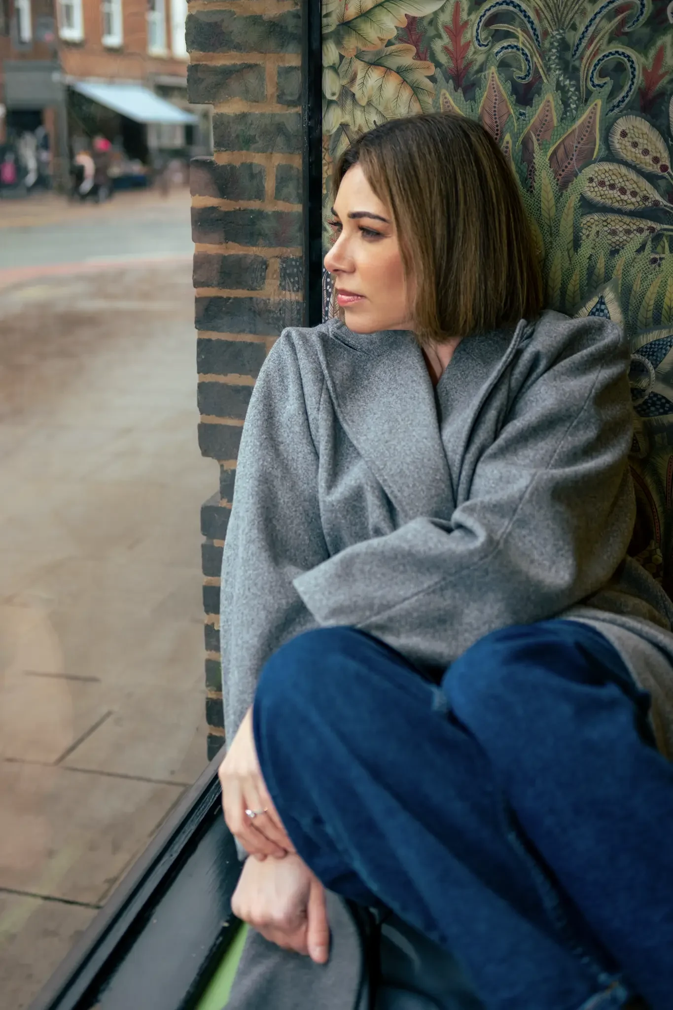 A woman with short brown hair sitting by a window, gazing outside with a thoughtful expression. She is wearing a gray coat and blue jeans, sitting with her knees drawn up. Outside the window, a blurry street scene is visible.