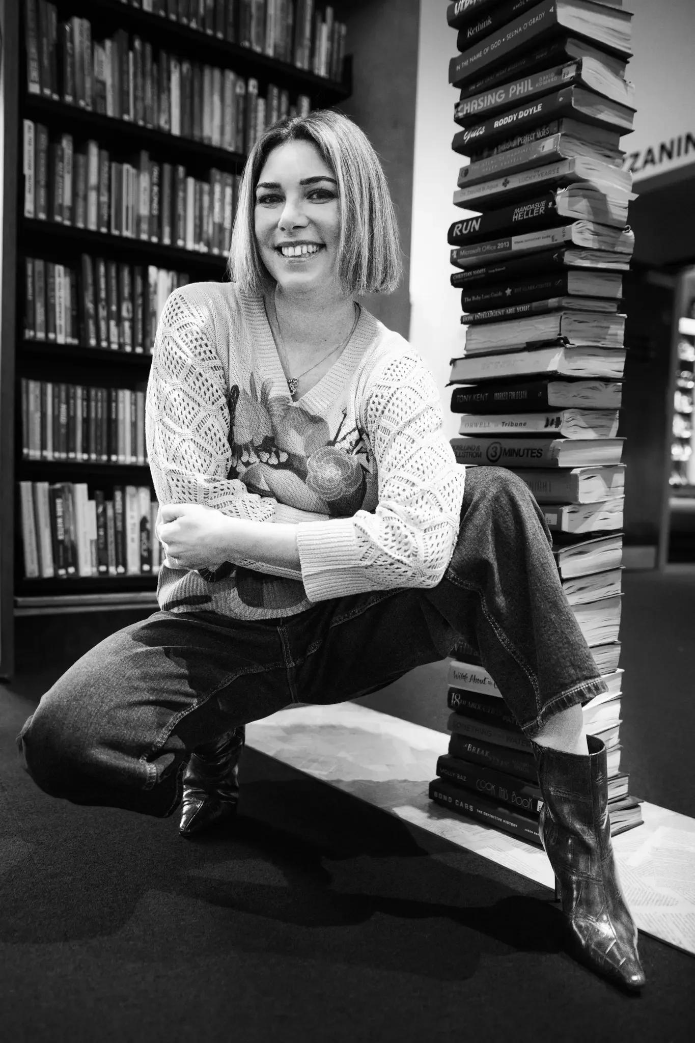 A woman with shoulder-length hair, smiling, crouching in a bookstore next to a tall stack of books, with bookshelves in the background.