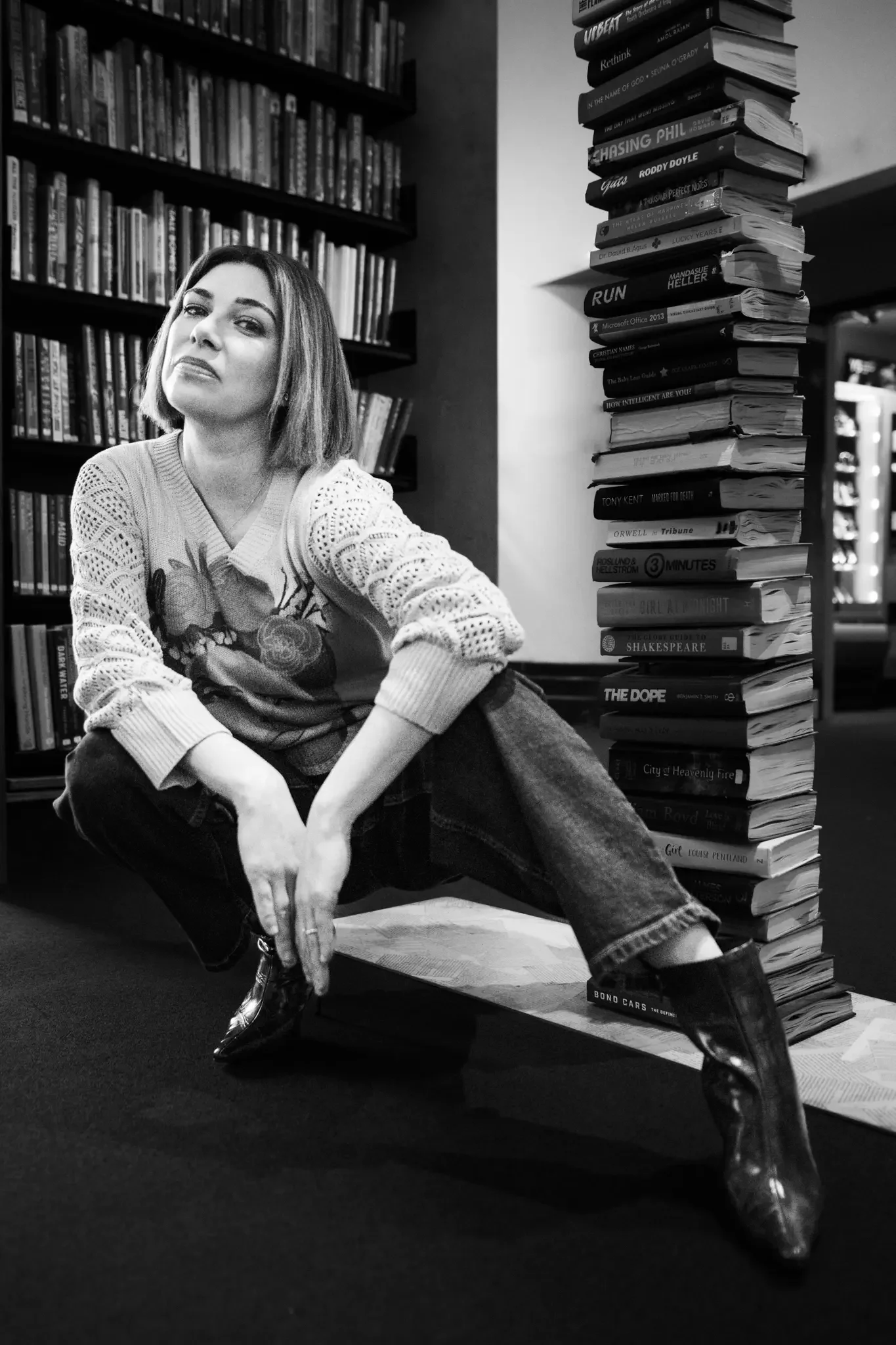 A woman with shoulder-length hair squatting in front of a tall stack of books indoors, with bookshelves in the background.