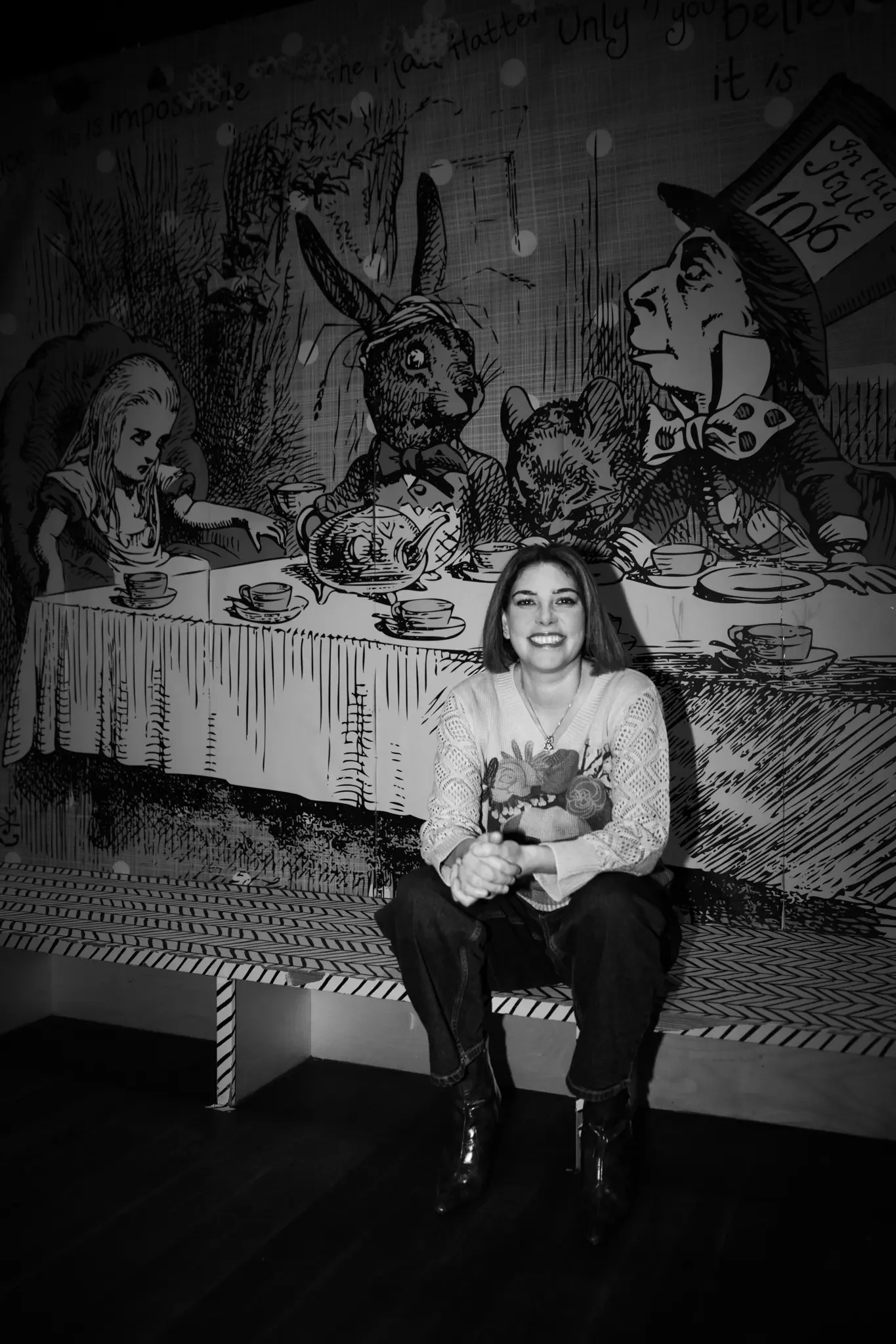 A woman with shoulder-length hair, smiling and sitting on a bench in front of a black and white mural depicting the Mad Hatter's tea party from Alice in Wonderland, featuring Alice, a rabbit, a cat, and a man with a large top hat.