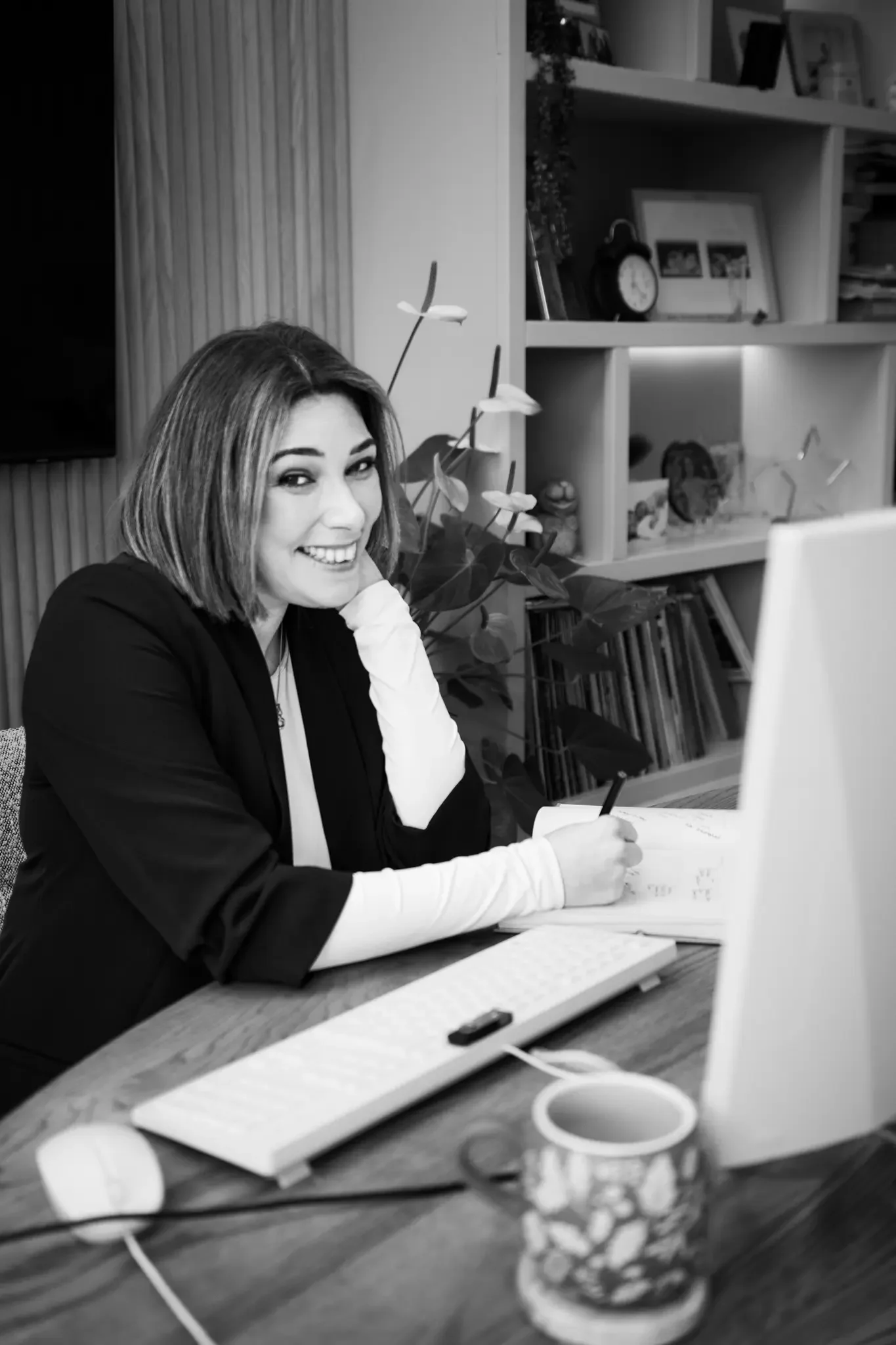 A woman with shoulder-length hair smiling and sitting at a desk with a computer, notebook, and coffee mug, in an office or home workspace.