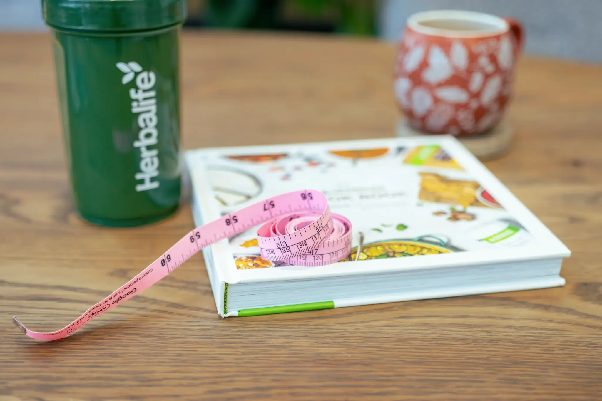 A green hydration bottle with 'Heartbar Life' written on it, a coiled pink measuring tape, a closed illustrated magazine or book, a red and white patterned mug, and a small wooden coaster on a wooden table.