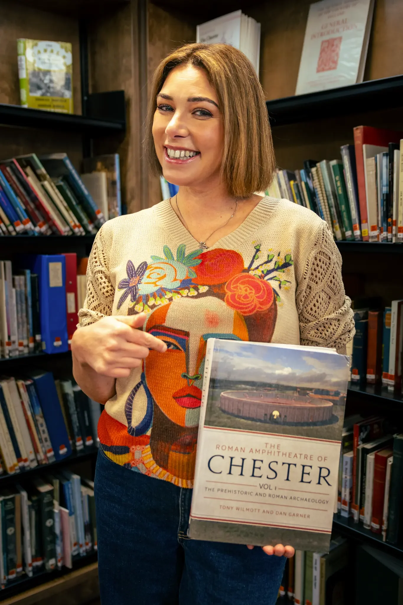 A woman standing in a library holding a book titled 'The Roman Amphitheatre of Chester Vol 1, The Prehistoric and Roman Archaeology' and smiling.