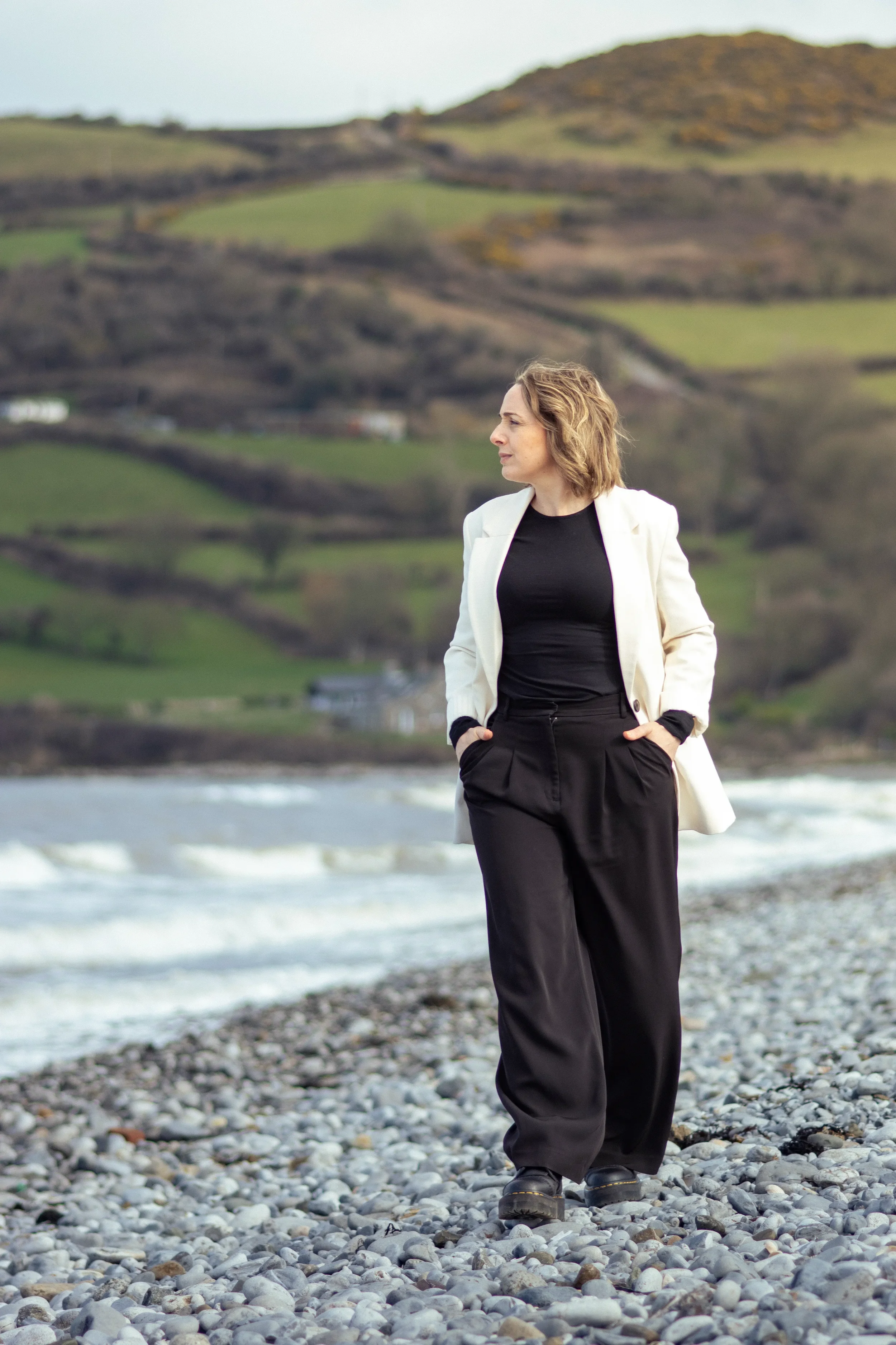 lady in a white blazer, black trousers and black DMs walks along a pebble beach.webp