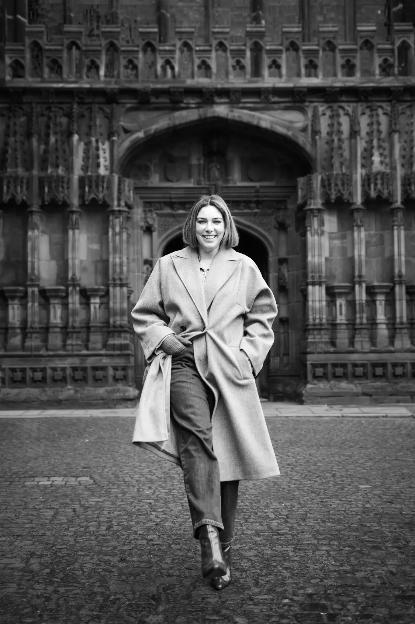 A woman walking in front of a historic gothic-style building, smiling, wearing a long coat, jeans, and ankle boots, in black and white.
