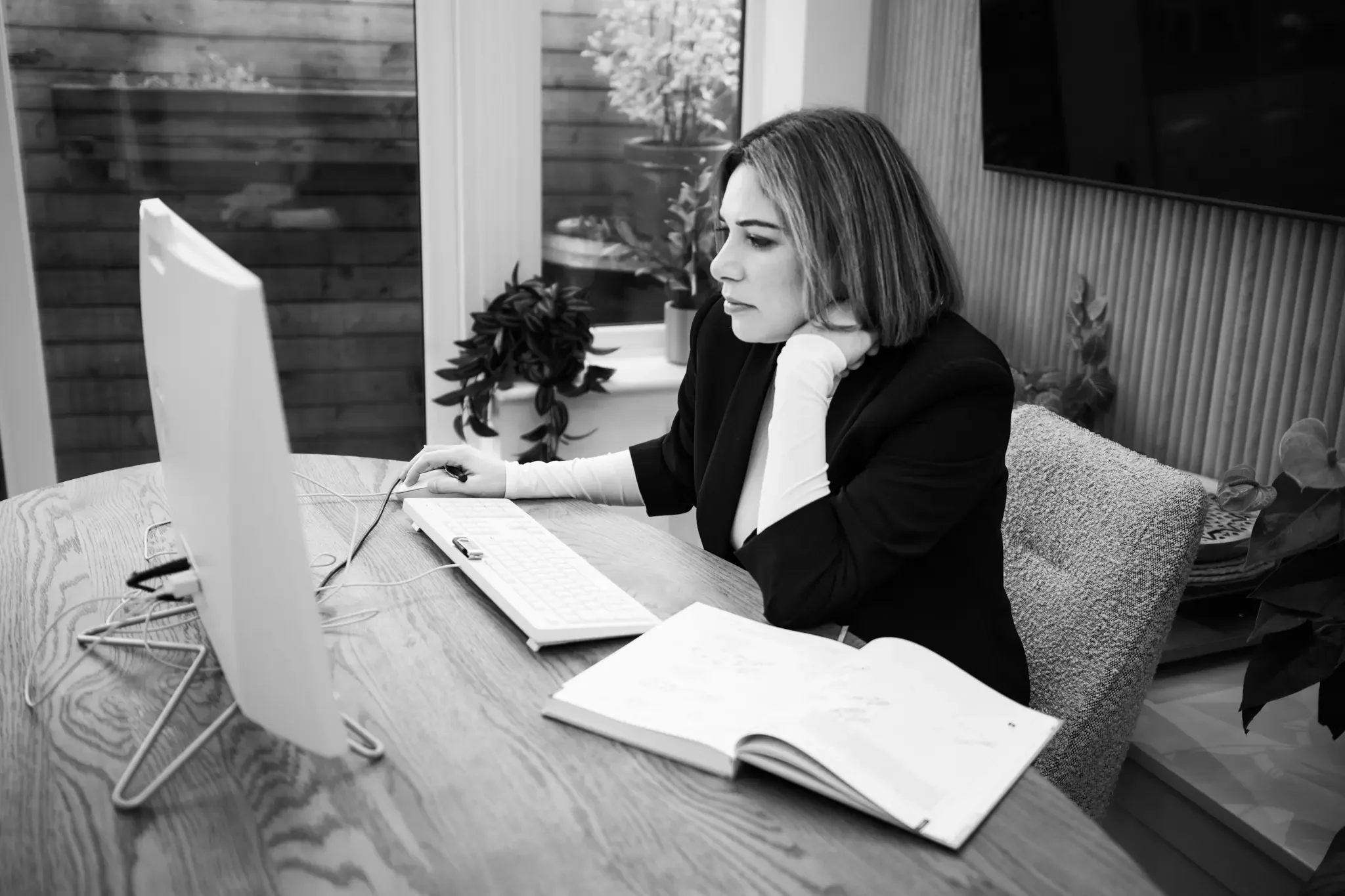 Black-and-white photo of a woman working at a desk, using a desktop computer, with an open book in front of her and a notebook in the background, in a room with large windows and plants.