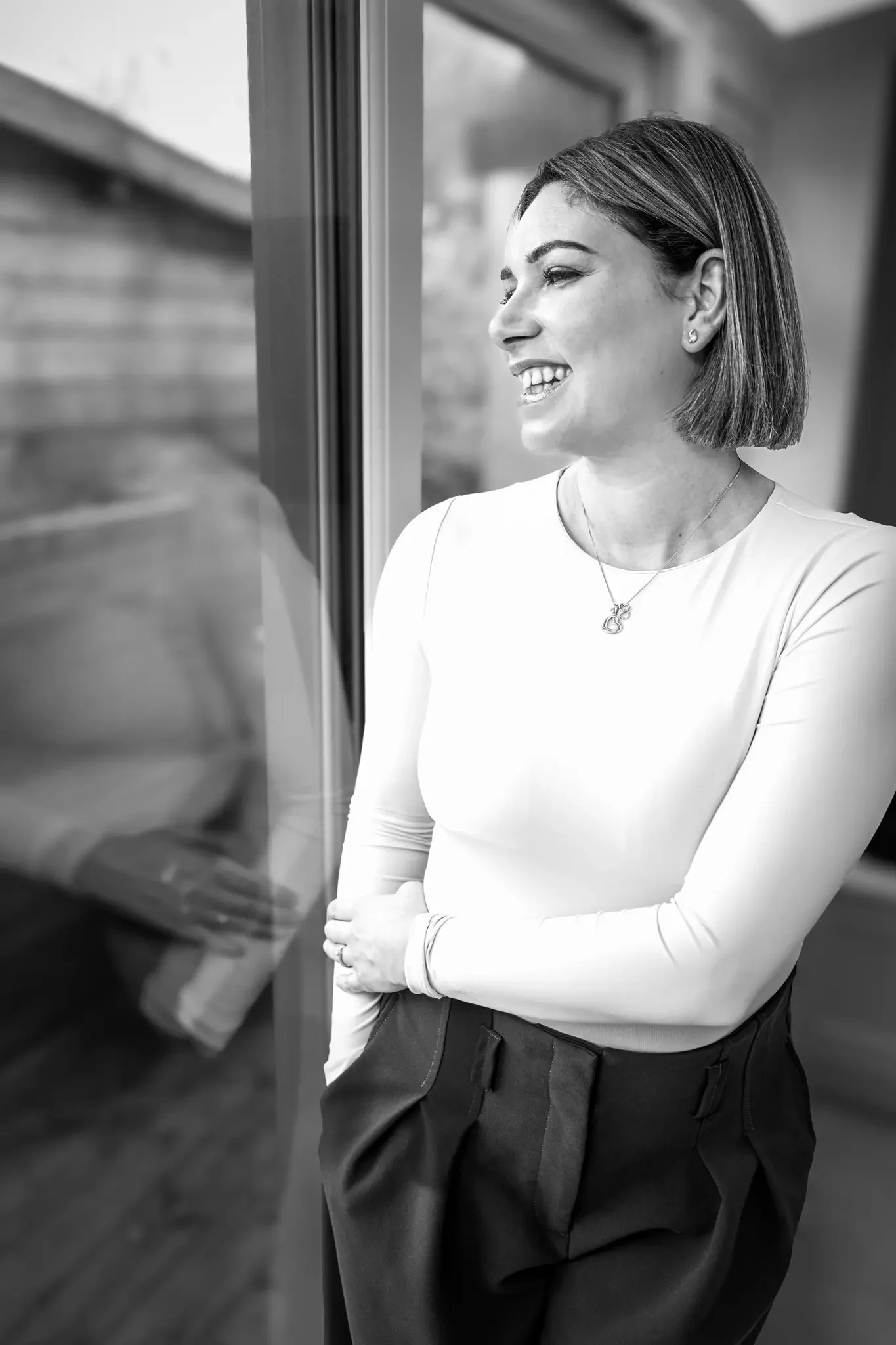 A woman with short hair and a necklace, smiling, standing with her arms crossed near a window in an indoor setting.