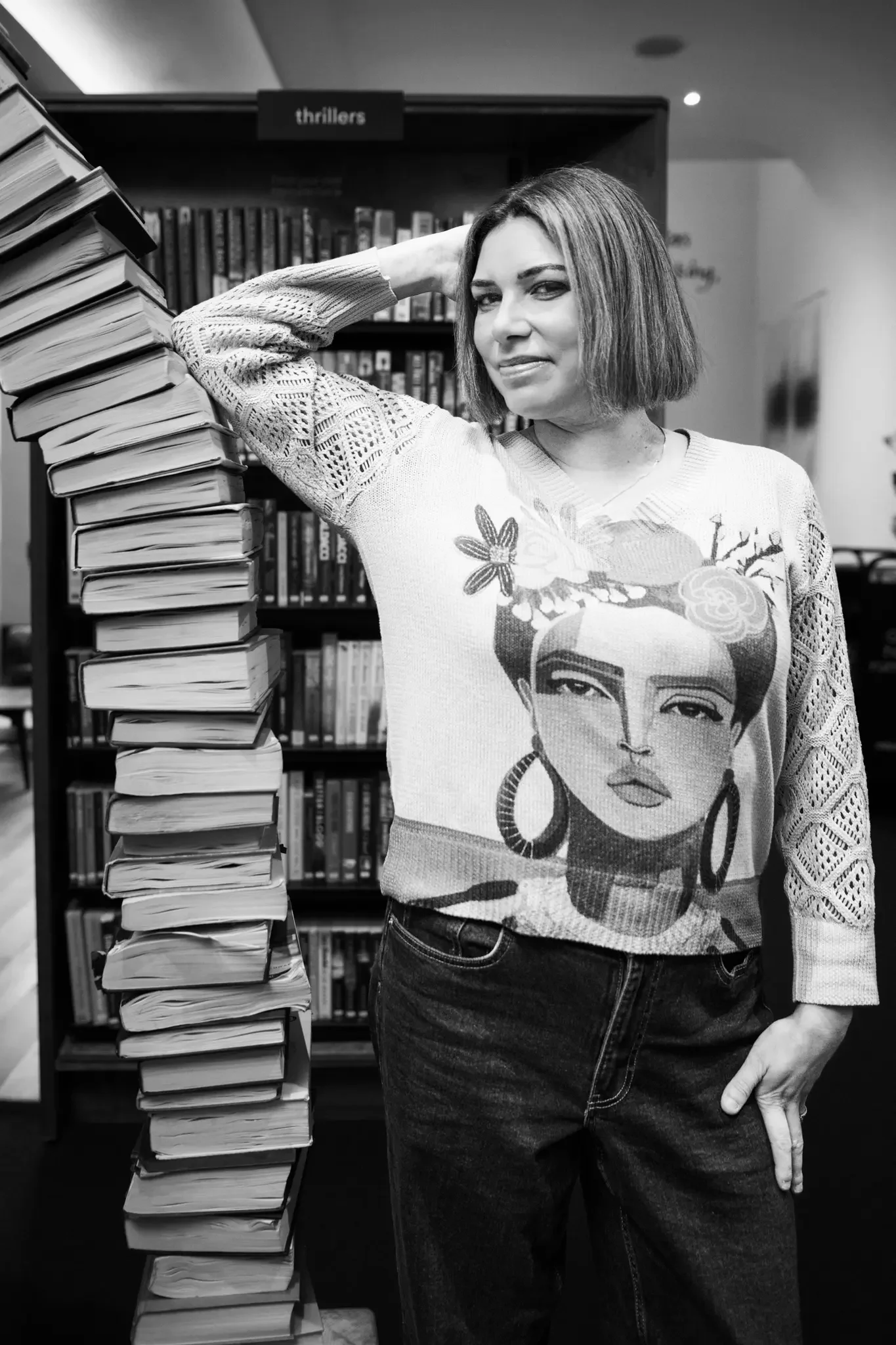 A woman with short hair standing in a library, leaning on a tall stack of books, with bookshelves behind her labeled 'thrillers'.