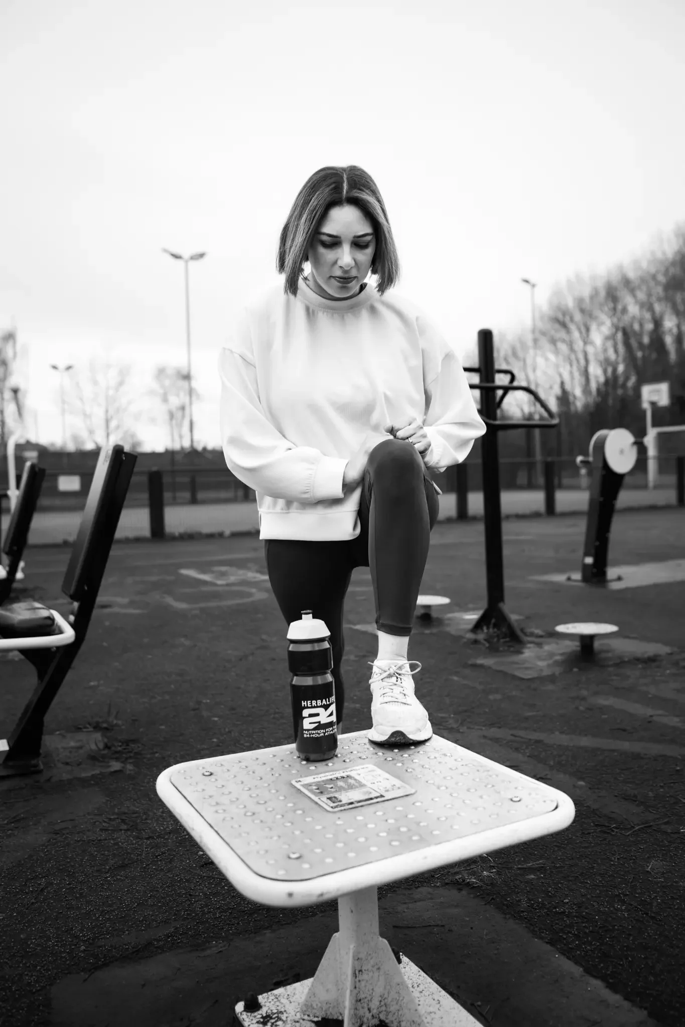 A woman at an outdoor gym is balancing on one leg on a small raised platform, holding her knee. She is dressed in workout clothes, including a sweatshirt, leggings, and sneakers. There is a water bottle and a snack bar on the platform. The background