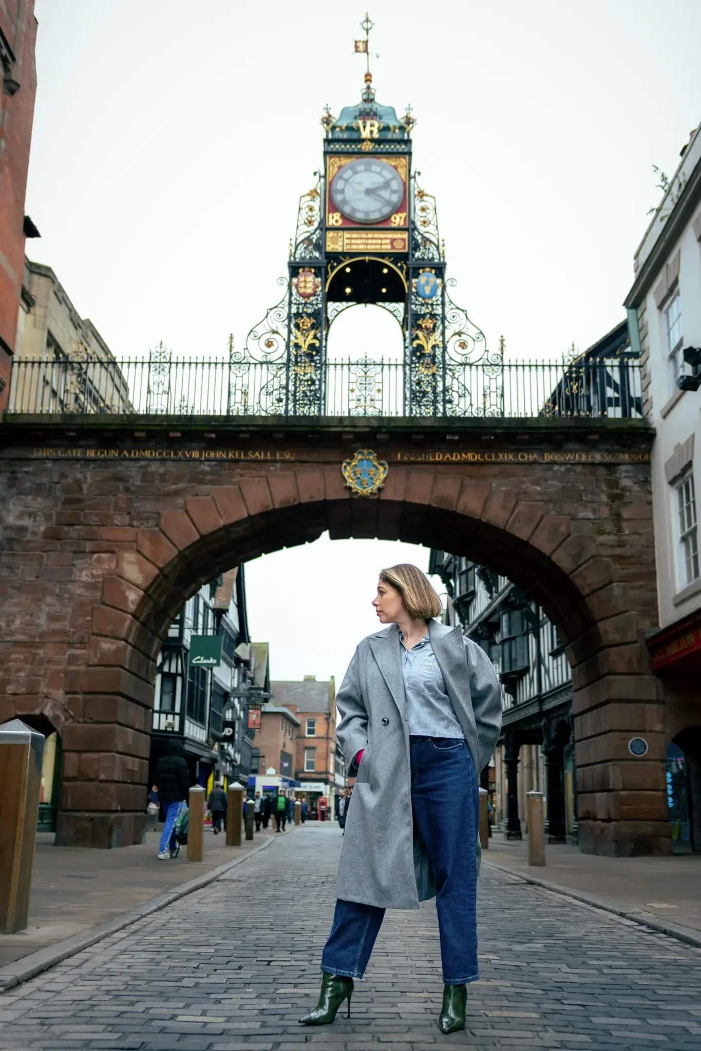 A woman standing on a cobblestone street under an archway with a decorative clock tower.