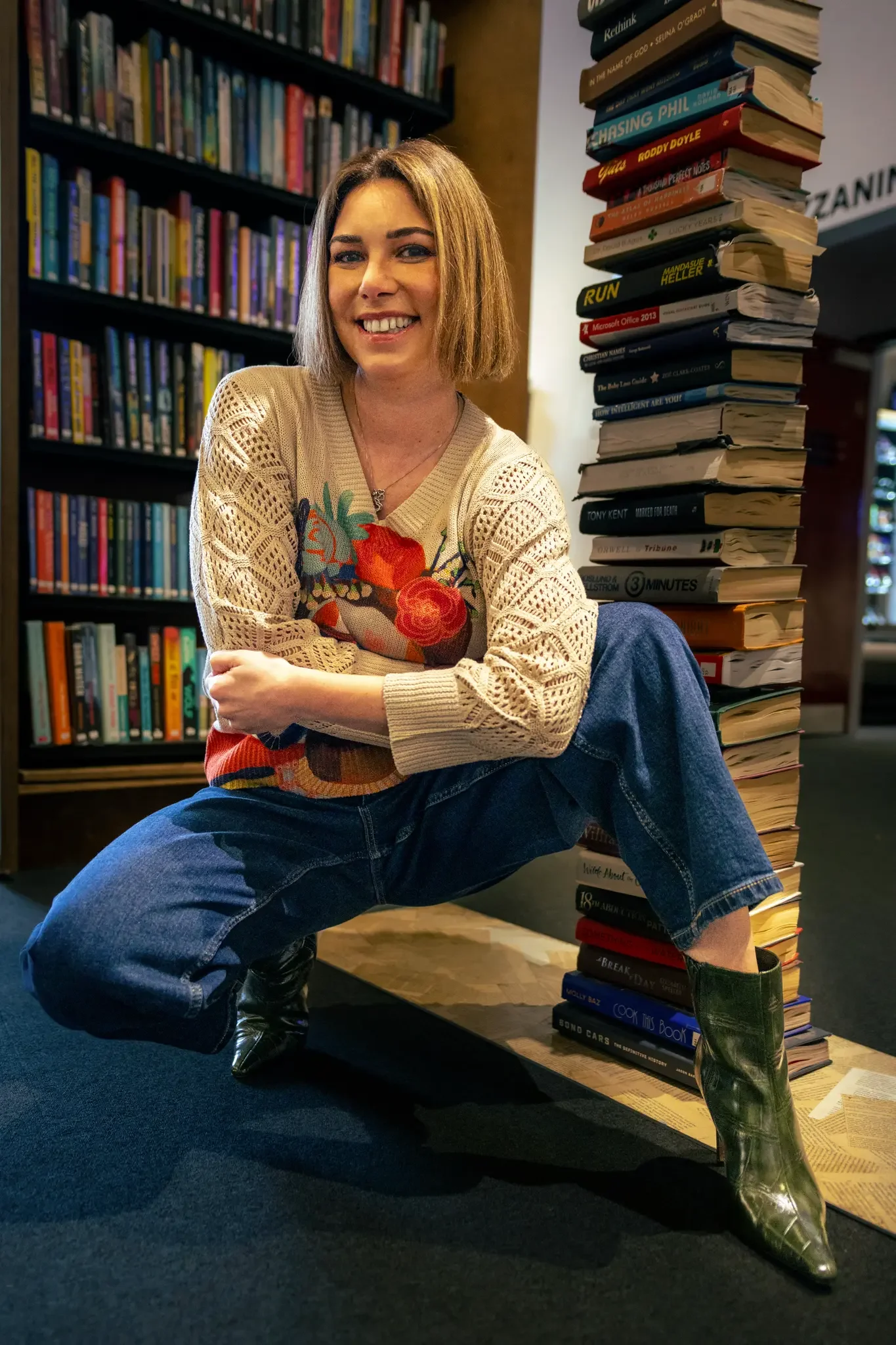 A woman with shoulder-length blonde hair and a big smile, sitting on the floor with one knee bent, next to a large stack of books. She is wearing a knit sweater with colorful floral embroidery, jeans, and shiny black boots. In the background, there a