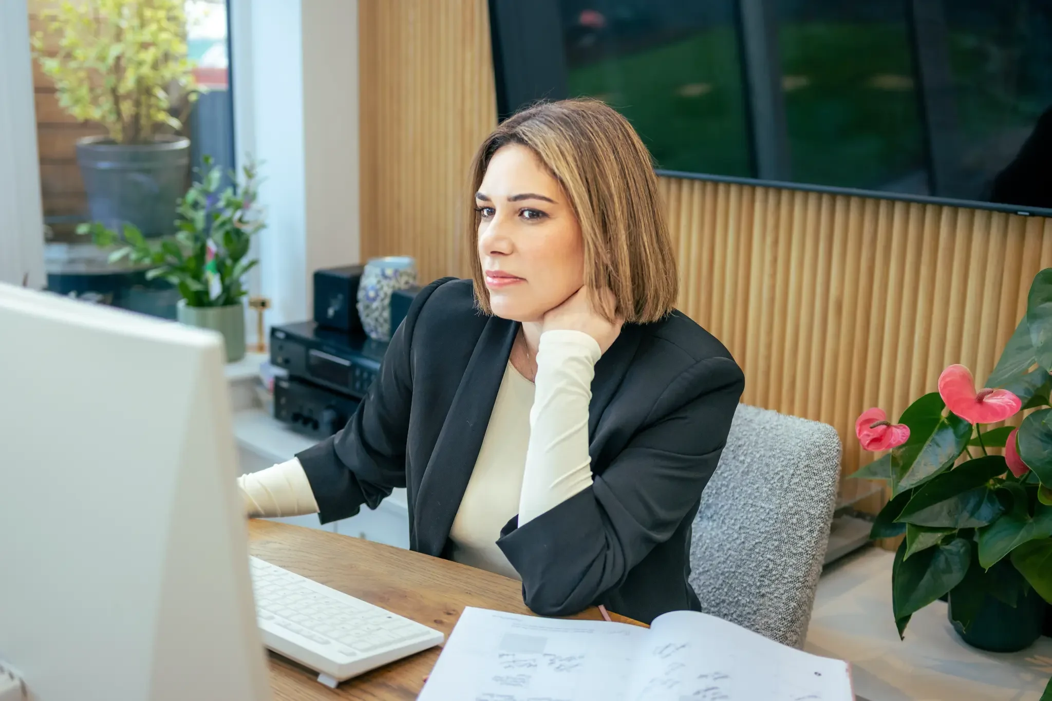 A woman sitting at a desk in an office, looking at a computer screen with a pensive expression, with pink flowering plants and indoor plants in the background.
