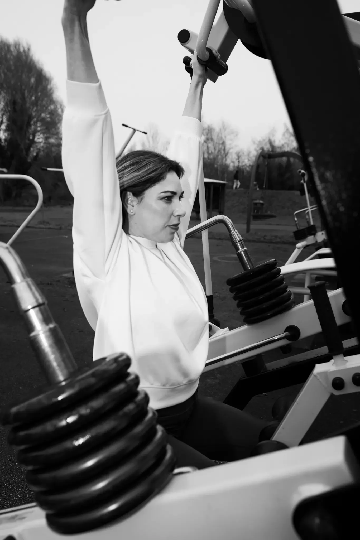 A woman in a white shirt is exercising on gym equipment outdoors, raising her arms above her head at a park.