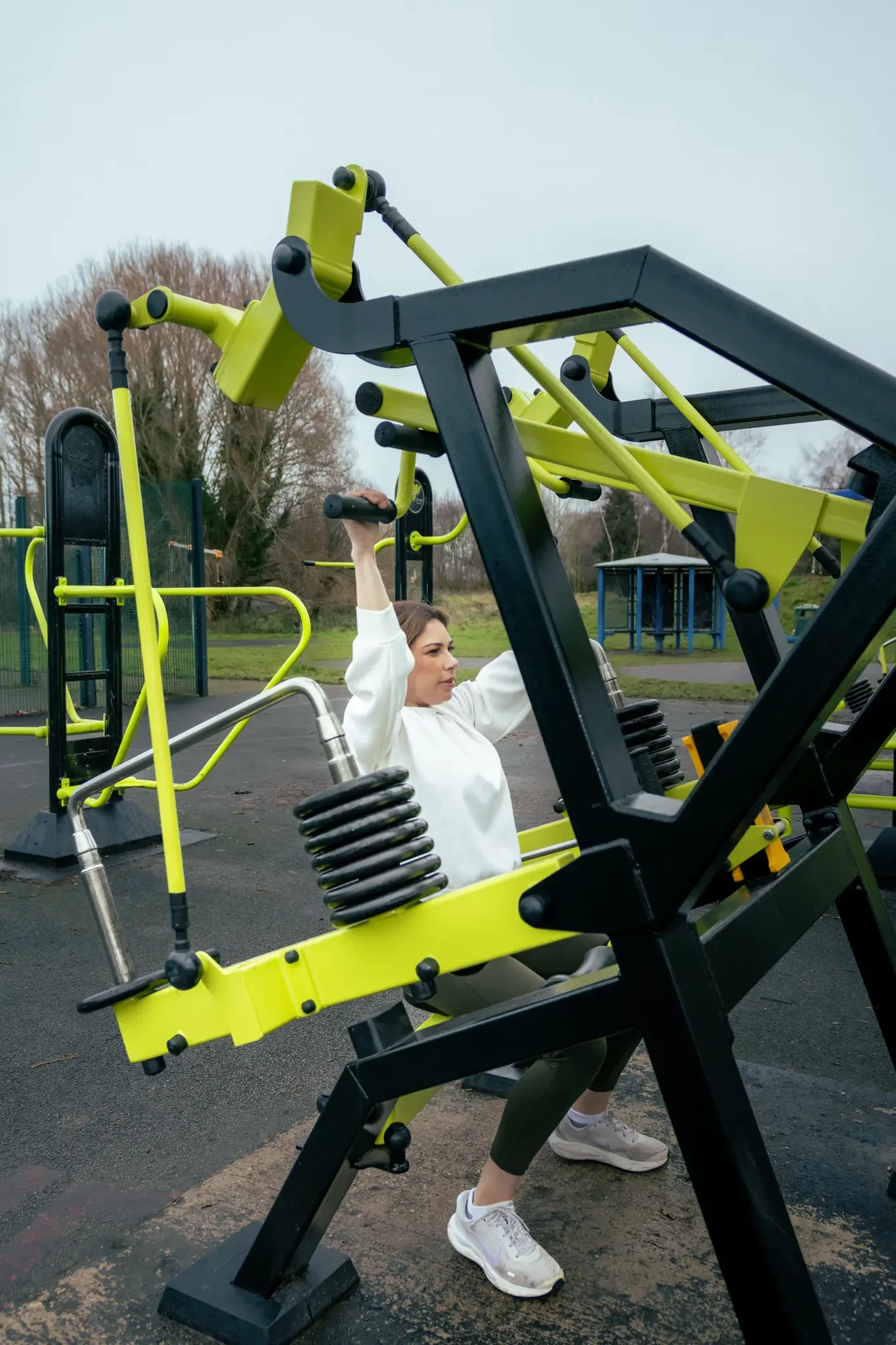 A woman in a white sweatshirt performing an exercise with outdoor gym equipment in a park, with trees and cloudy sky in the background.