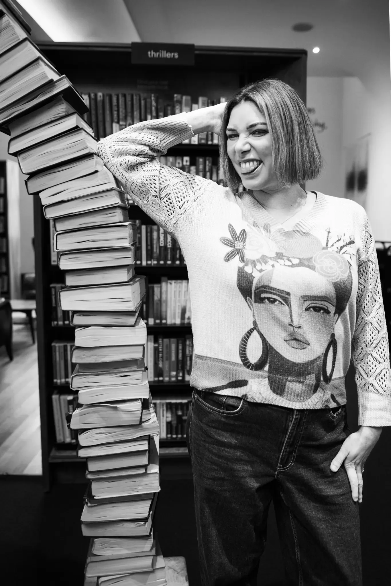 A woman with shoulder-length hair making a playful grimace, standing next to a tall stack of books in a library.