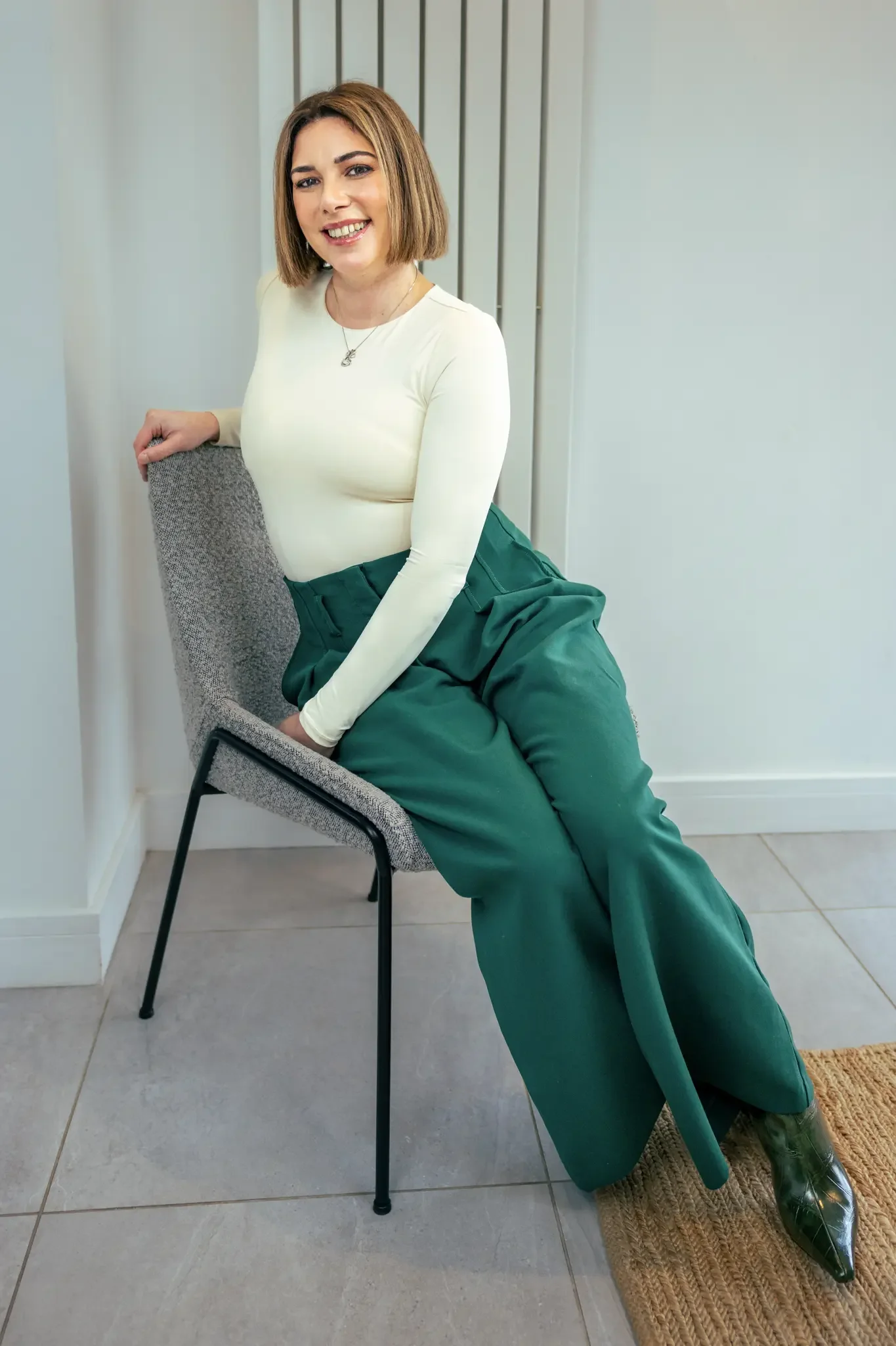 A woman with shoulder-length brown hair, wearing a cream long-sleeve top, green high-waisted pants, and green boots, seated on a gray chair, smiling at the camera in a modern room with gray tiled flooring and white walls.