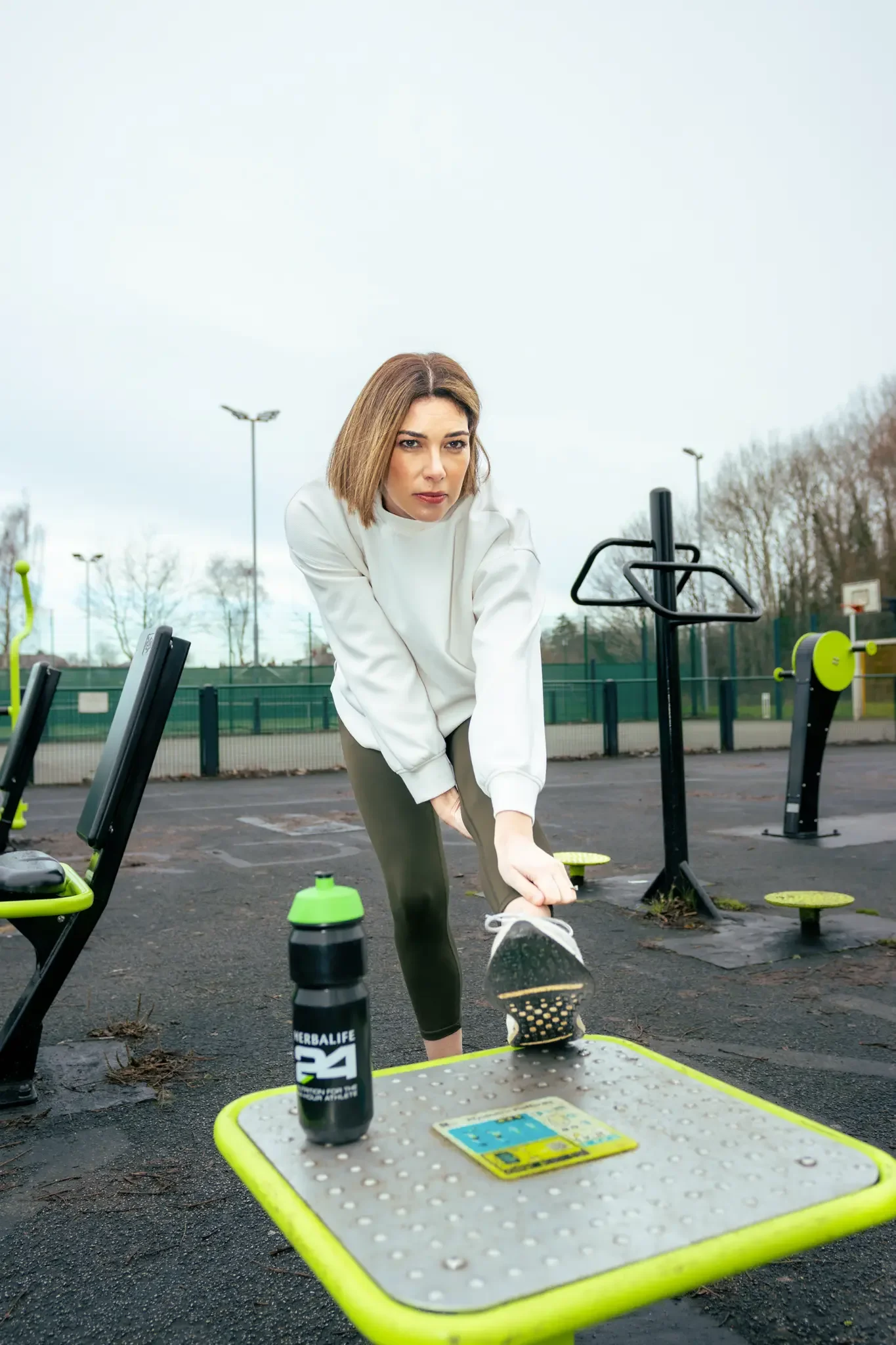 Woman in workout clothes stretching outdoors at a gym, with exercise equipment and a water bottle on a nearby table.