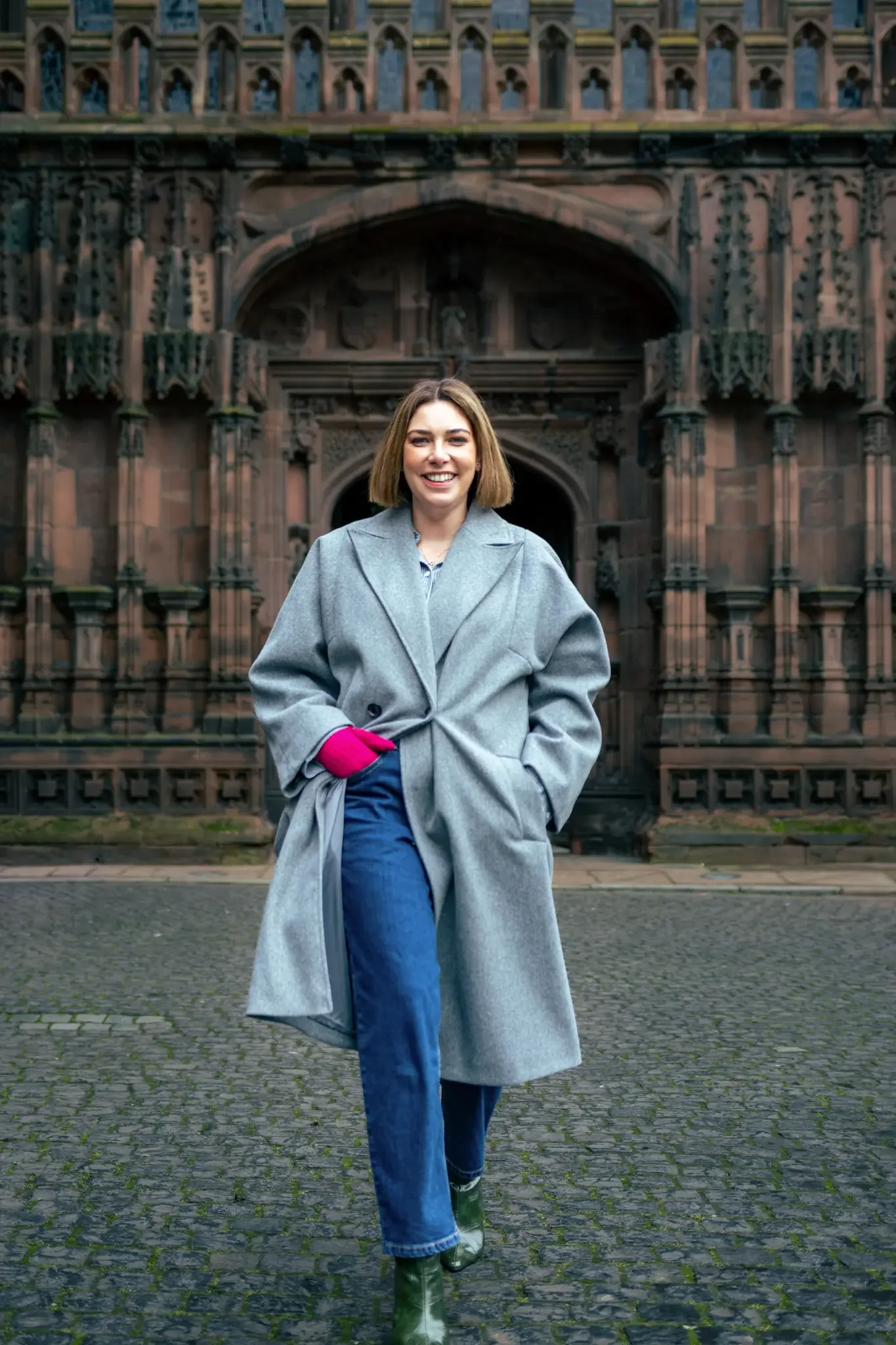 A woman smiling and walking on a cobblestone street in front of a historic Gothic cathedral, wearing a gray coat, blue jeans, green boots, and pink gloves.