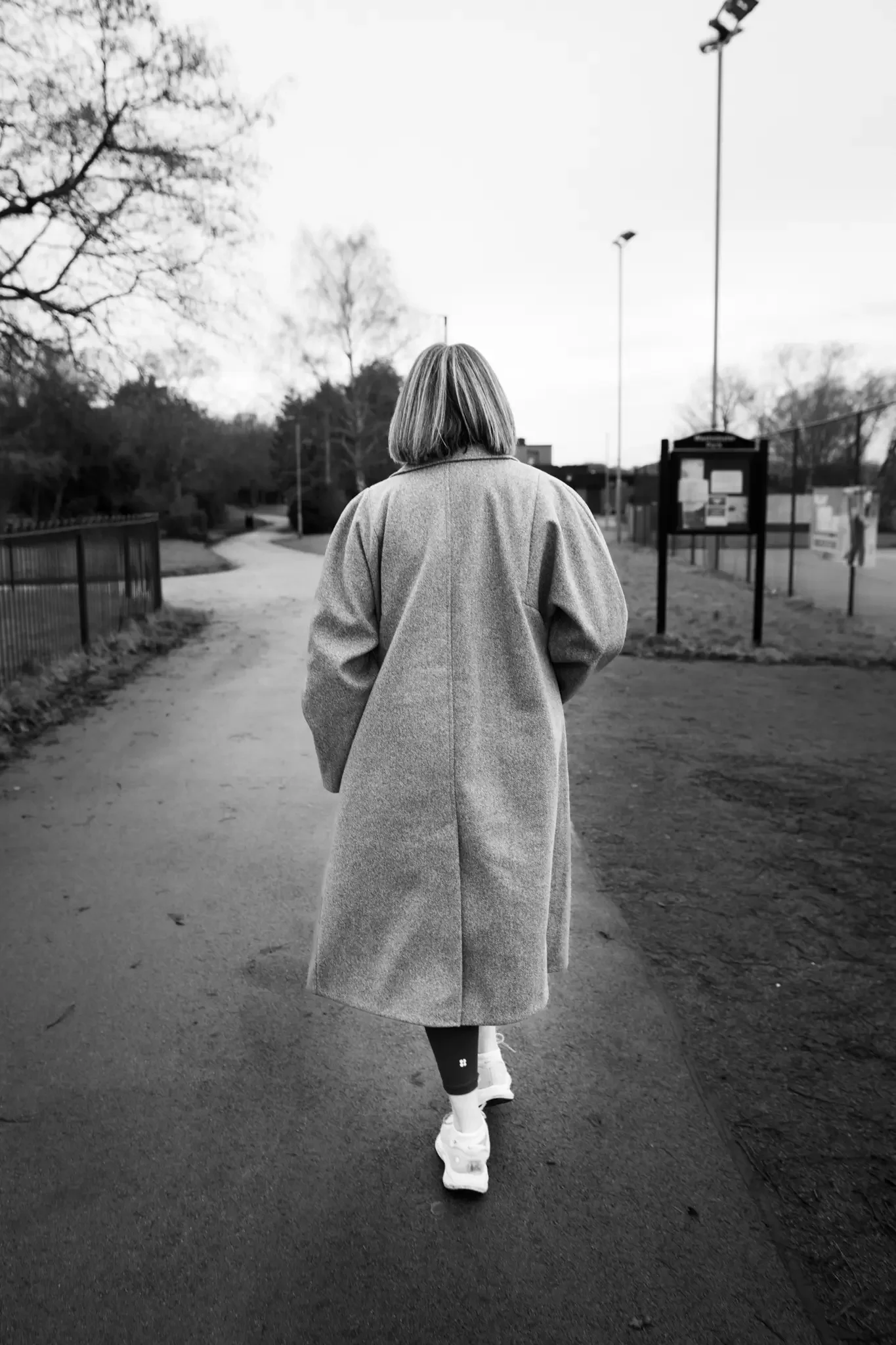 A woman with shoulder-length hair, dressed in a long coat and sneakers, walking away on a paved path in a park or outdoor area, with trees, fences, and informational signs in the background, captured in black and white.