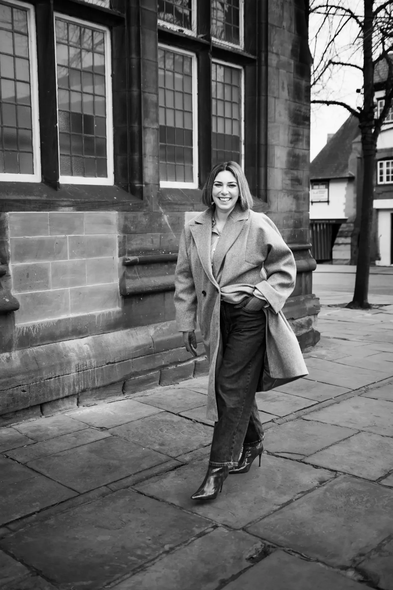 A woman smiling and walking on a city sidewalk near a historical building, wearing a long coat, high-heeled boots, and jeans, in black and white.