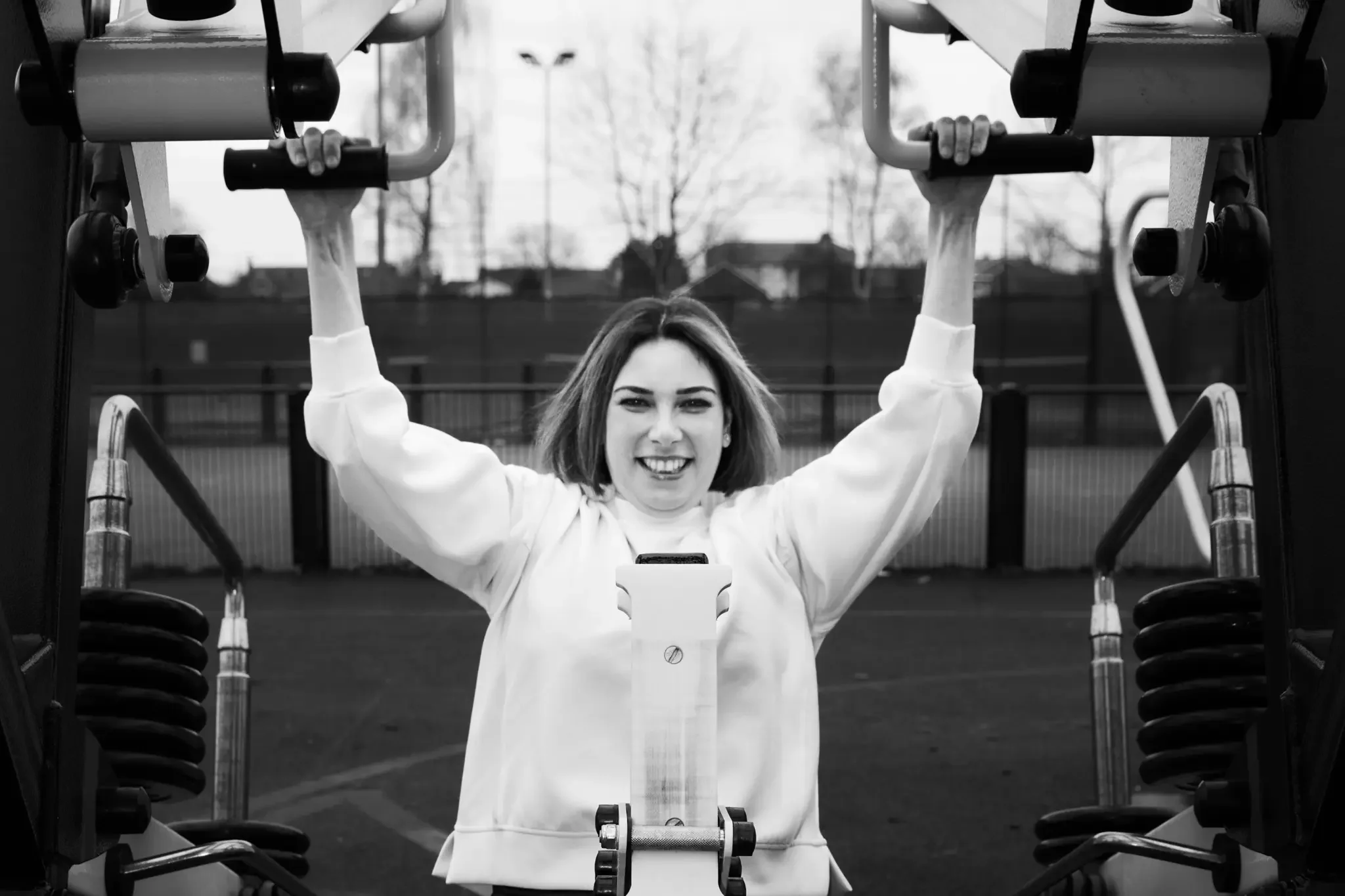 A woman with short hair and a white sweatshirt smiling and hanging from a horizontal bar at a gym or outdoor workout area.