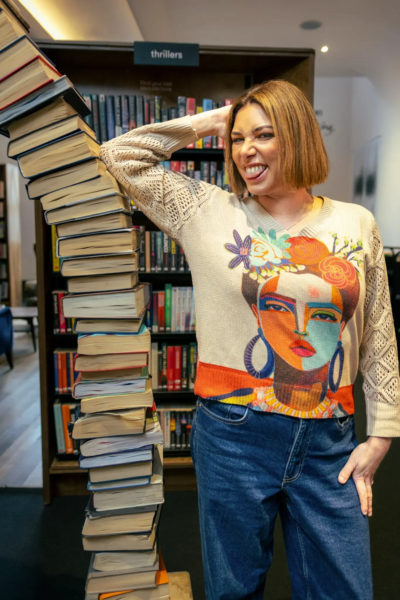 A woman with shoulder-length reddish hair making a playful face, standing next to a tall stack of books in a library or bookstore, behind a bookshelf labeled 'thrillers.' She is wearing a colorful sweater with a stylized woman's face and blue jeans.