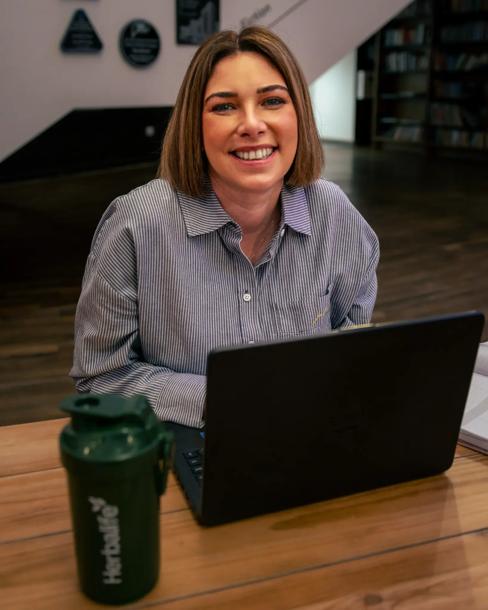 Smiling woman sitting at a wooden table with a laptop and a green water bottle in front of her, in a room with bookshelves and wall decor.
