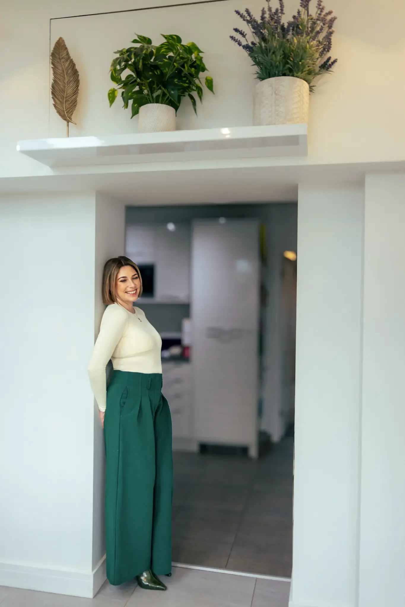 A woman with shoulder-length brown hair smiling and standing with her back against a white wall in a modern interior space, wearing a cream-colored top, green high-waisted pants, and shiny green shoes, with potted plants and decorative items on a she