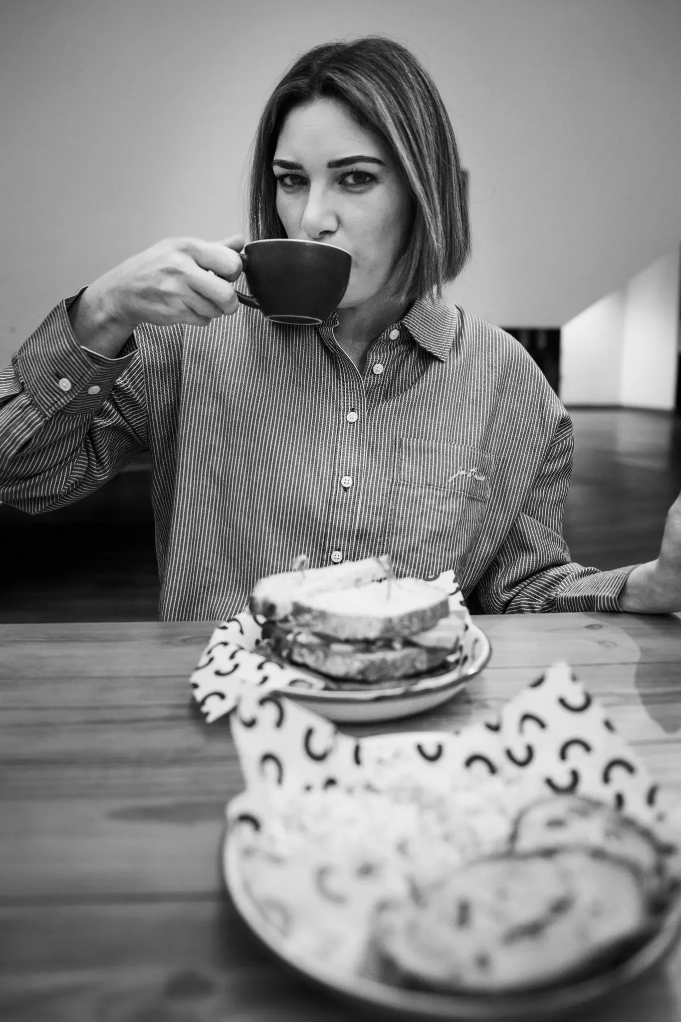 A woman with shoulder-length hair drinking from a cup at a table with plates of sandwiches and cookies.