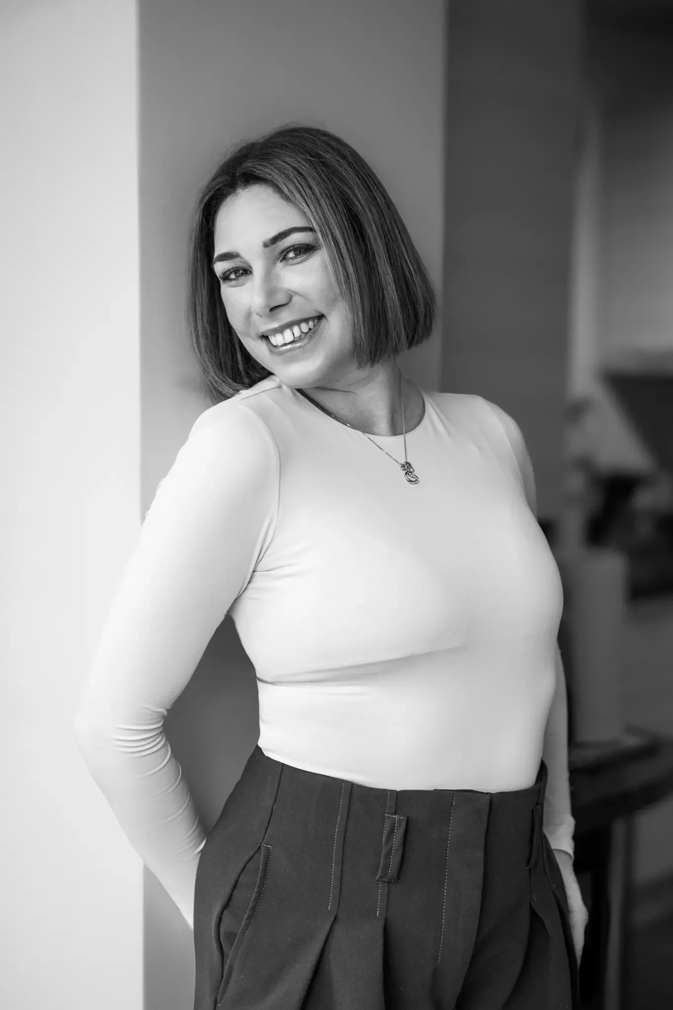 A woman with shoulder-length hair smiling and leaning against a wall, wearing a long-sleeved top and high-waisted pants in a black-and-white photo.
