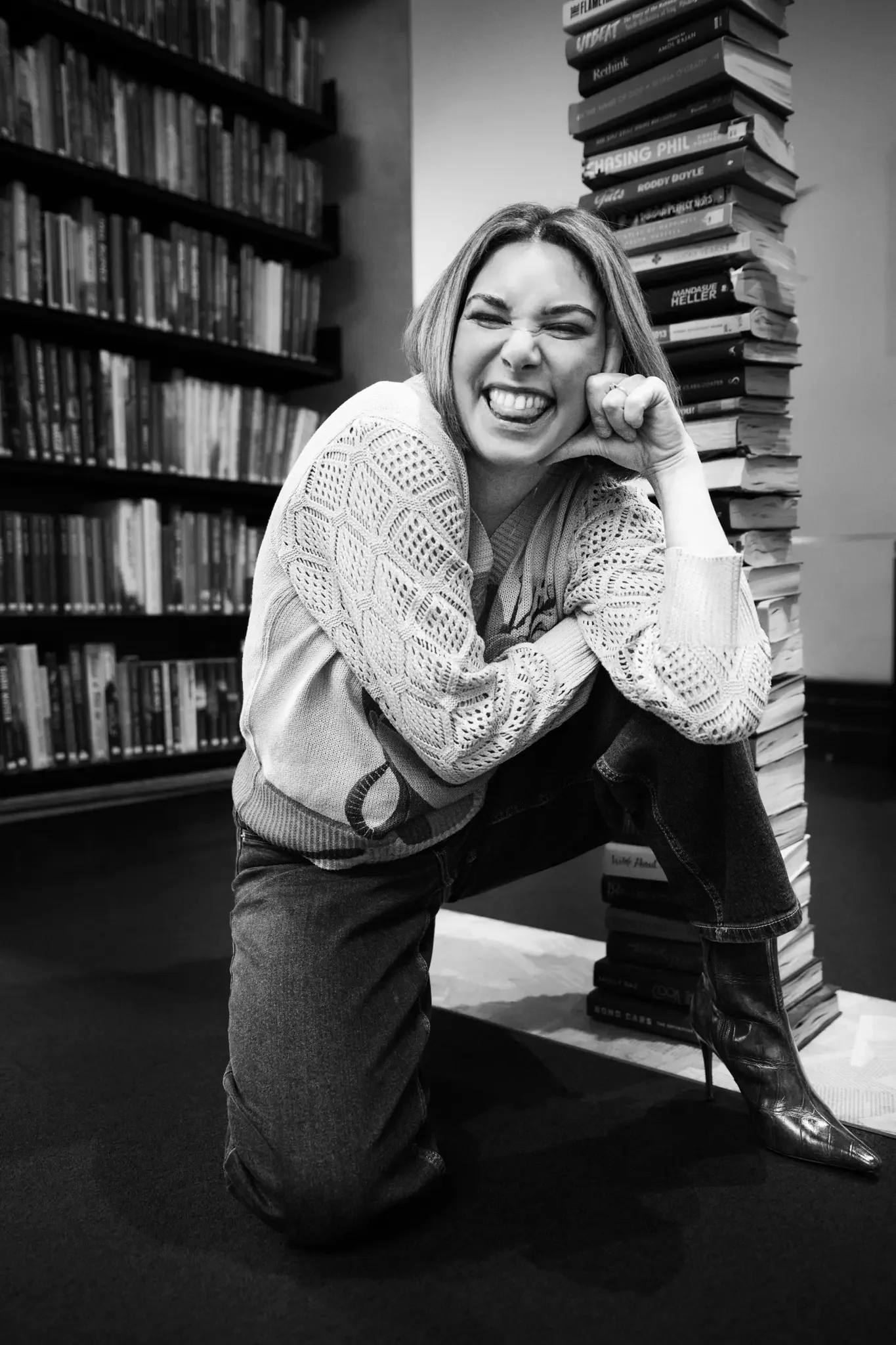 A woman with shoulder-length hair squatting down in a library, smiling and making a playful face, with a large stack of books beside her and more bookshelves in the background.