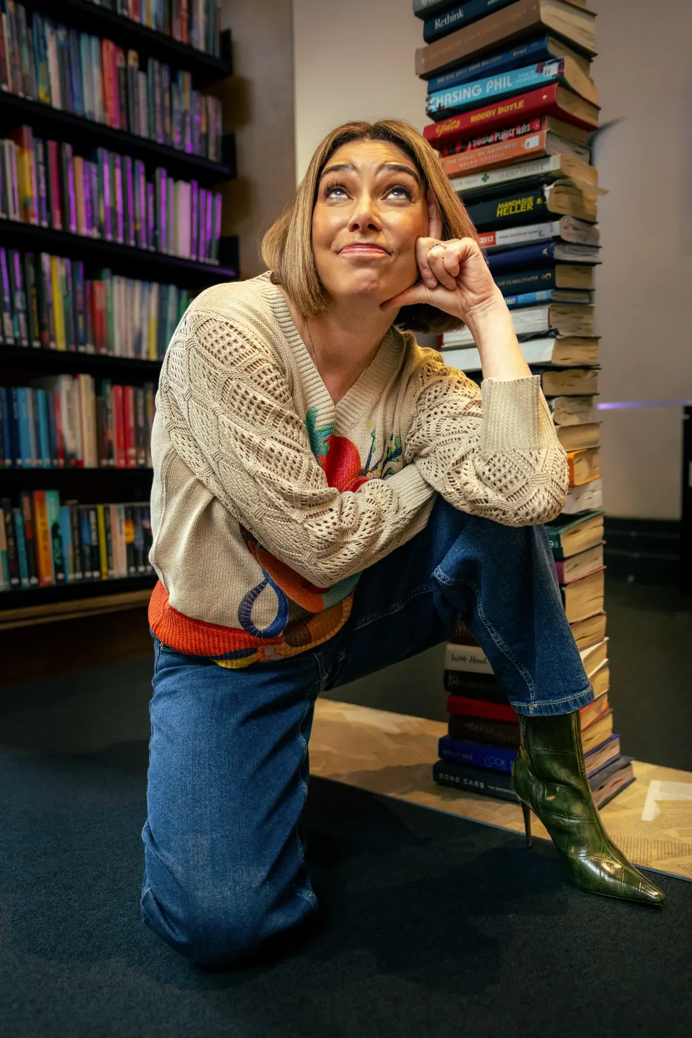 A woman with shoulder-length hair, wearing a patterned sweater, blue jeans, and green high-heeled boots, sitting on her knee in a library surrounded by stacked books and bookshelves filled with colorful books.